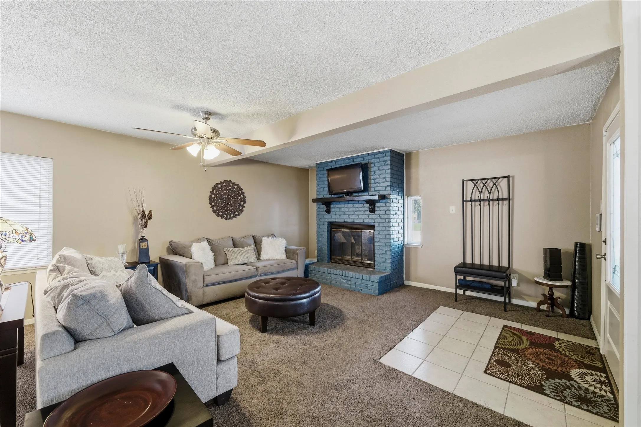 Living Room features a textured ceiling with ceiling fan and light fixture. Painted brick fireplace with raised hearth and wood mantel anchors the space. Windows provide natural light throughout. Carpet flooring extends throughout.