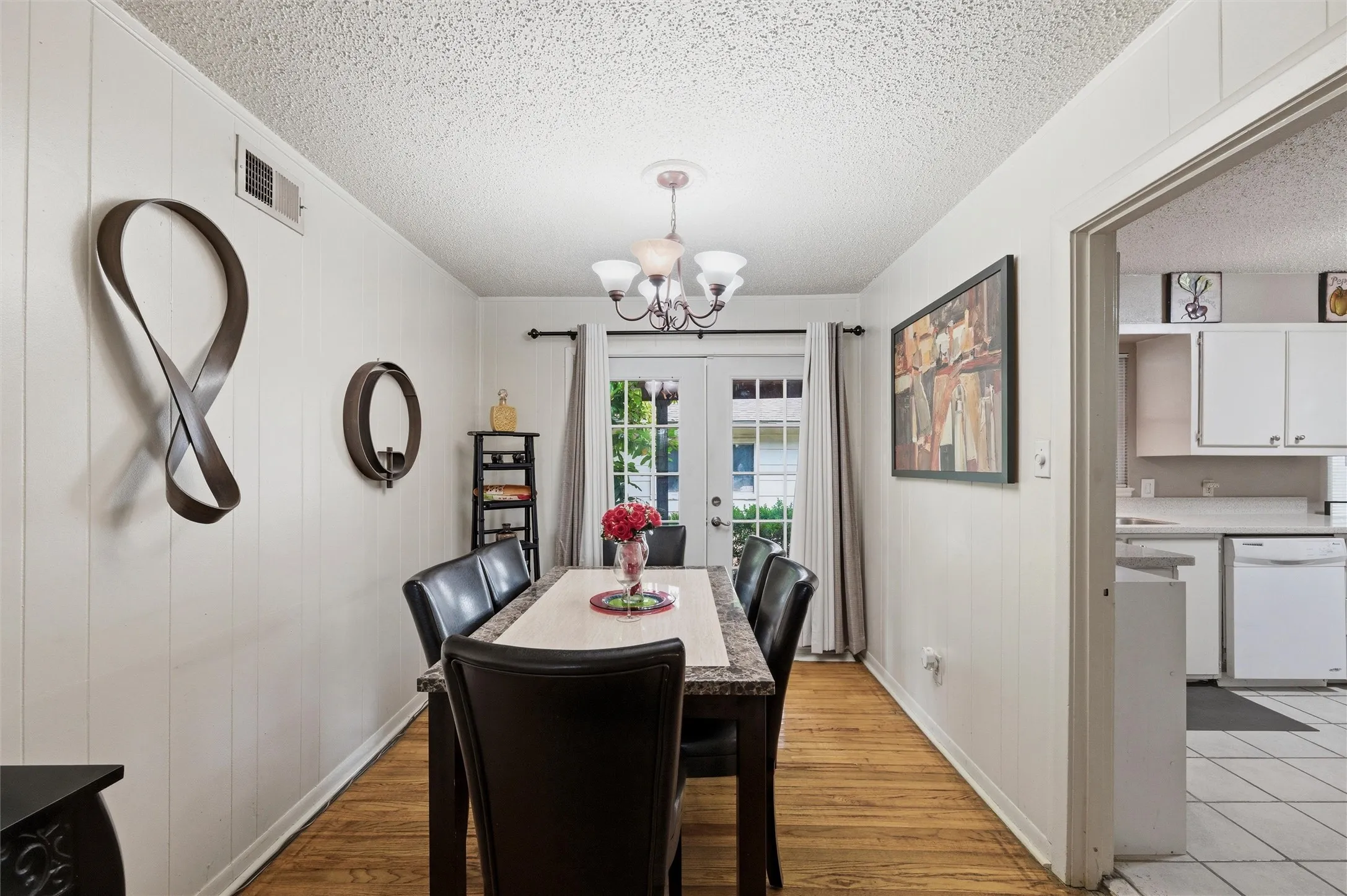 Dining Area features a textured ceiling with chandelier light fixture. Vertical board wall paneling accents the space. French doors provide natural light and exterior access. Open layout connects directly to the kitchen. Hardwood flooring extends throughout.