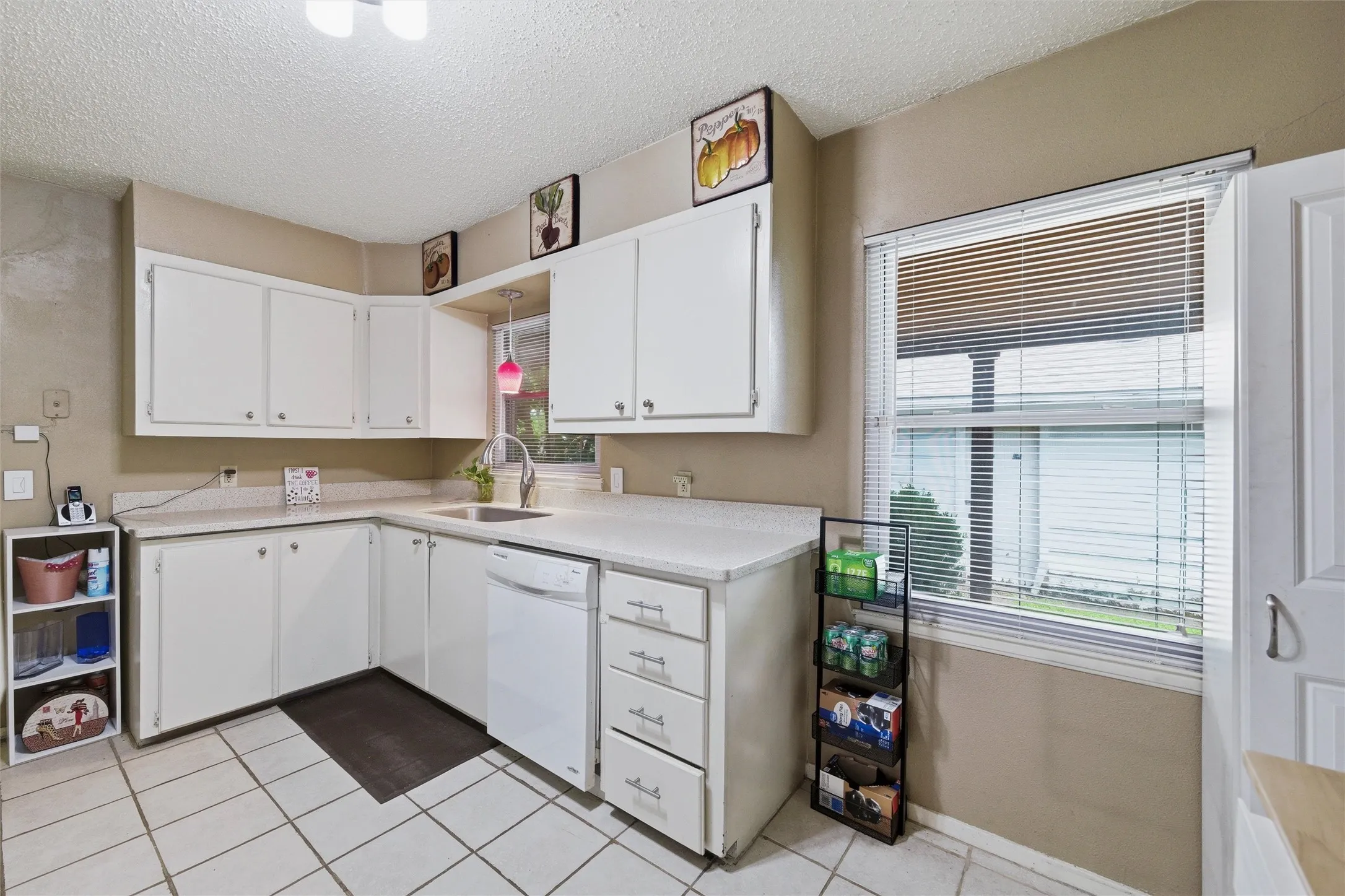 Kitchen features a textured ceiling with flush-mount light fixture and pendant lighting above the sink. White raised-panel cabinetry extends throughout with laminate countertops and single-basin sink. Window above the sink provides natural light. Tile flooring extends throughout.