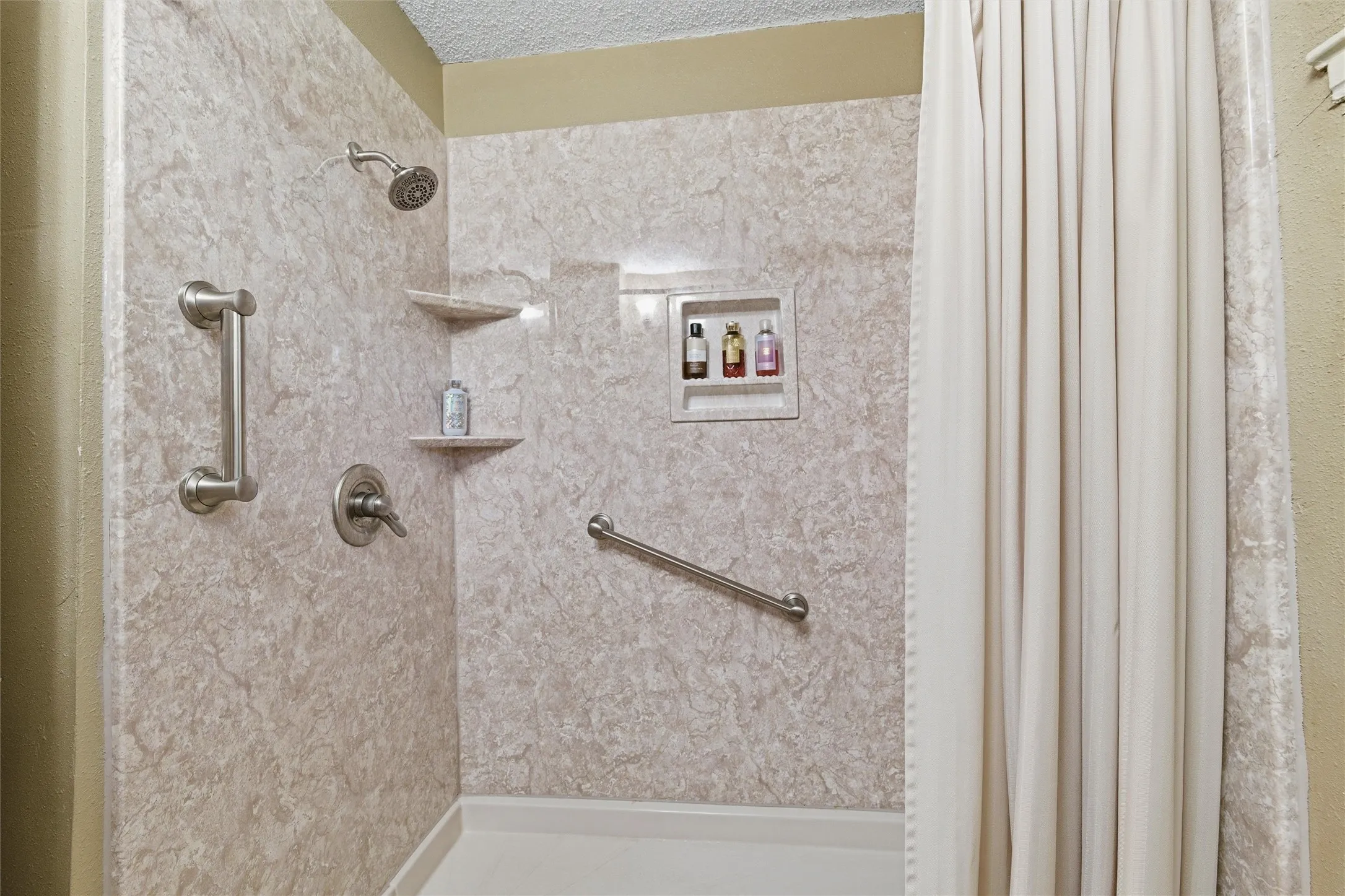 Bathroom features a textured ceiling with vanity lighting. Walk-in shower includes full-height surround, built-in niche shelving, corner shelving, and grab bars. Window provides natural light. Tile flooring extends throughout.
