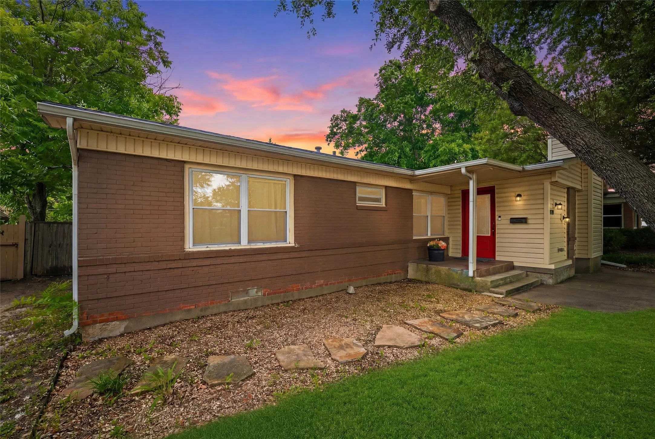 Front Exterior features a composition roof with gutter system, painted brick construction, and vertical board siding. Windows are positioned along the front elevation. Covered entry includes stepped concrete access and wall-mounted exterior lighting. Concrete driveway extends along the side of the property.