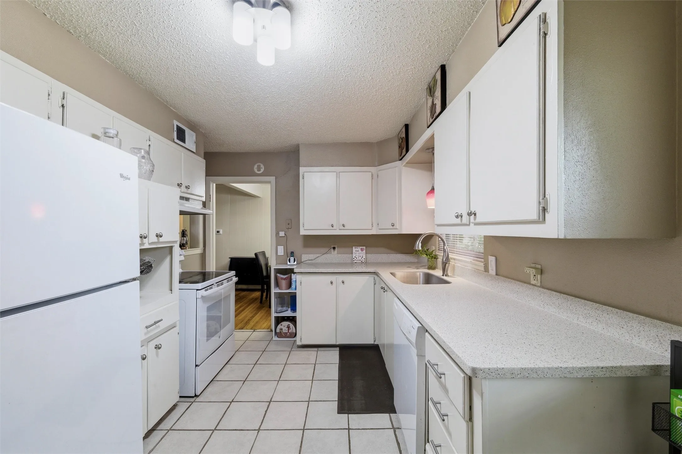 Kitchen features a textured ceiling with flush-mount light fixture. White raised-panel cabinetry extends throughout with laminate countertops and single-basin sink. Appliances include an electric range, dishwasher, and refrigerator. Open layout connects directly to the dining area. Tile flooring extends throughout.
