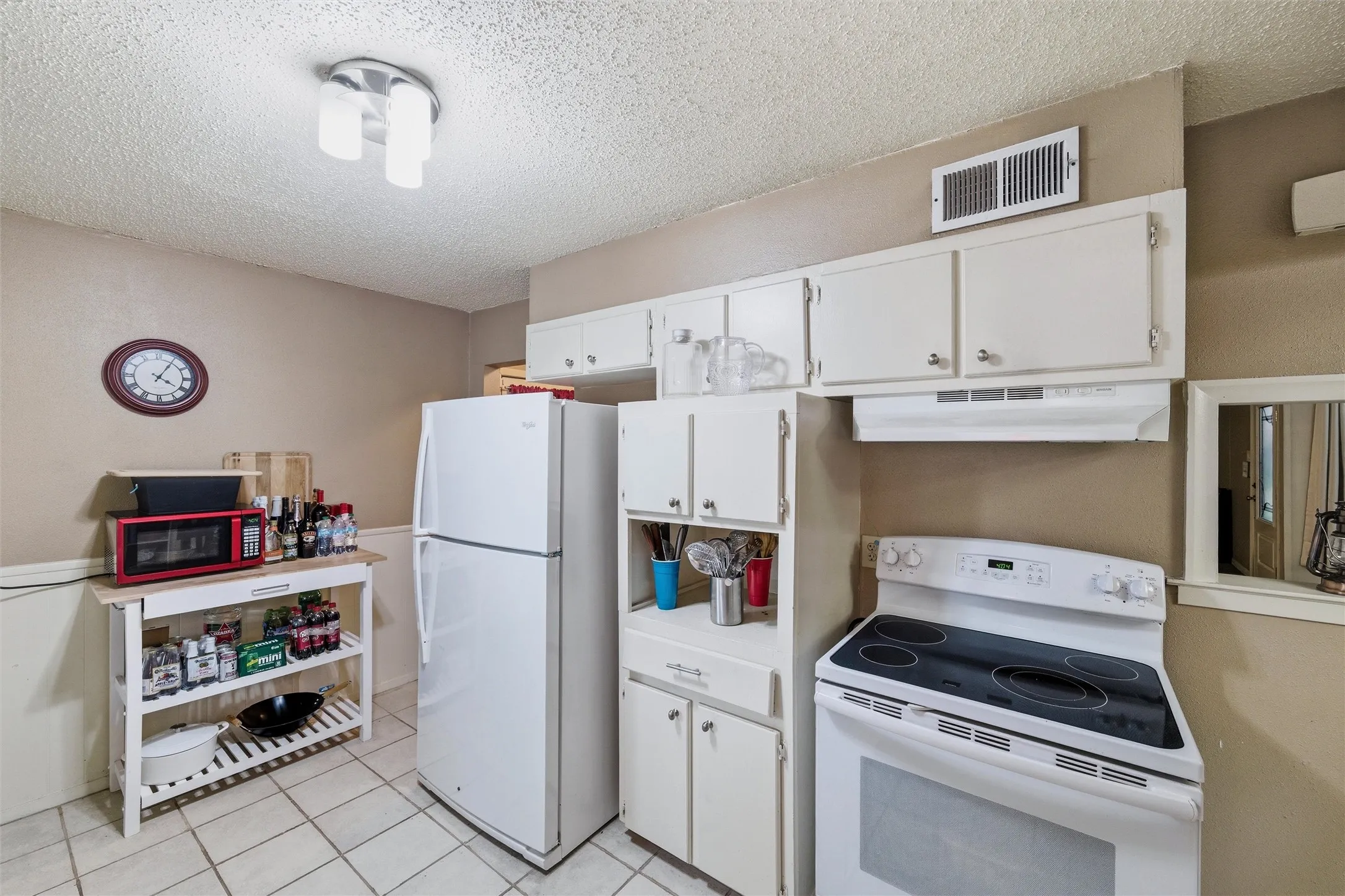 Kitchen features a textured ceiling with flush-mount light fixture. White raised-panel cabinetry provides storage throughout the space. Appliances include an electric range, vent hood, and refrigerator. Tile flooring extends throughout.