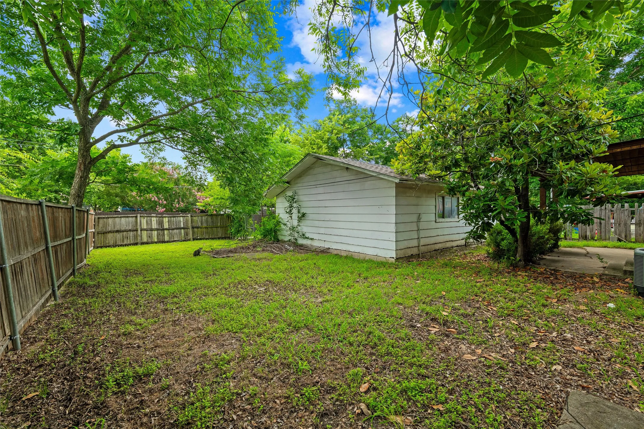 Backyard features mature trees providing shade throughout the property. Detached storage building includes a pitched roof, siding exterior, and side window. Wood fencing encloses the backyard. Grass lawn extends throughout.