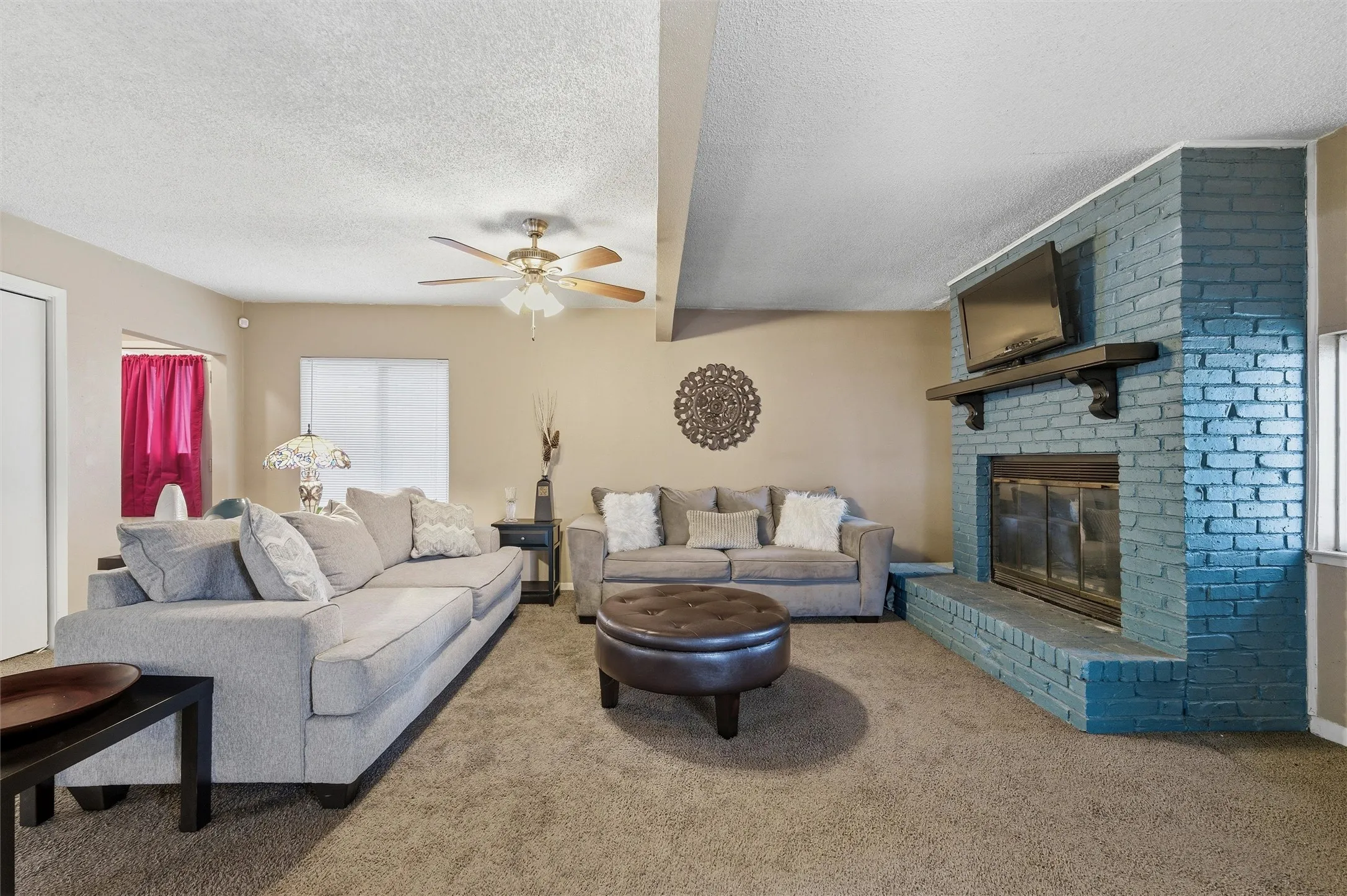 Living Room features a textured ceiling with ceiling fan and light fixture. Painted brick fireplace with raised hearth and wood mantel accents the room. Windows provide natural light. Open layout connects directly to the dining area. Carpet flooring extends throughout.