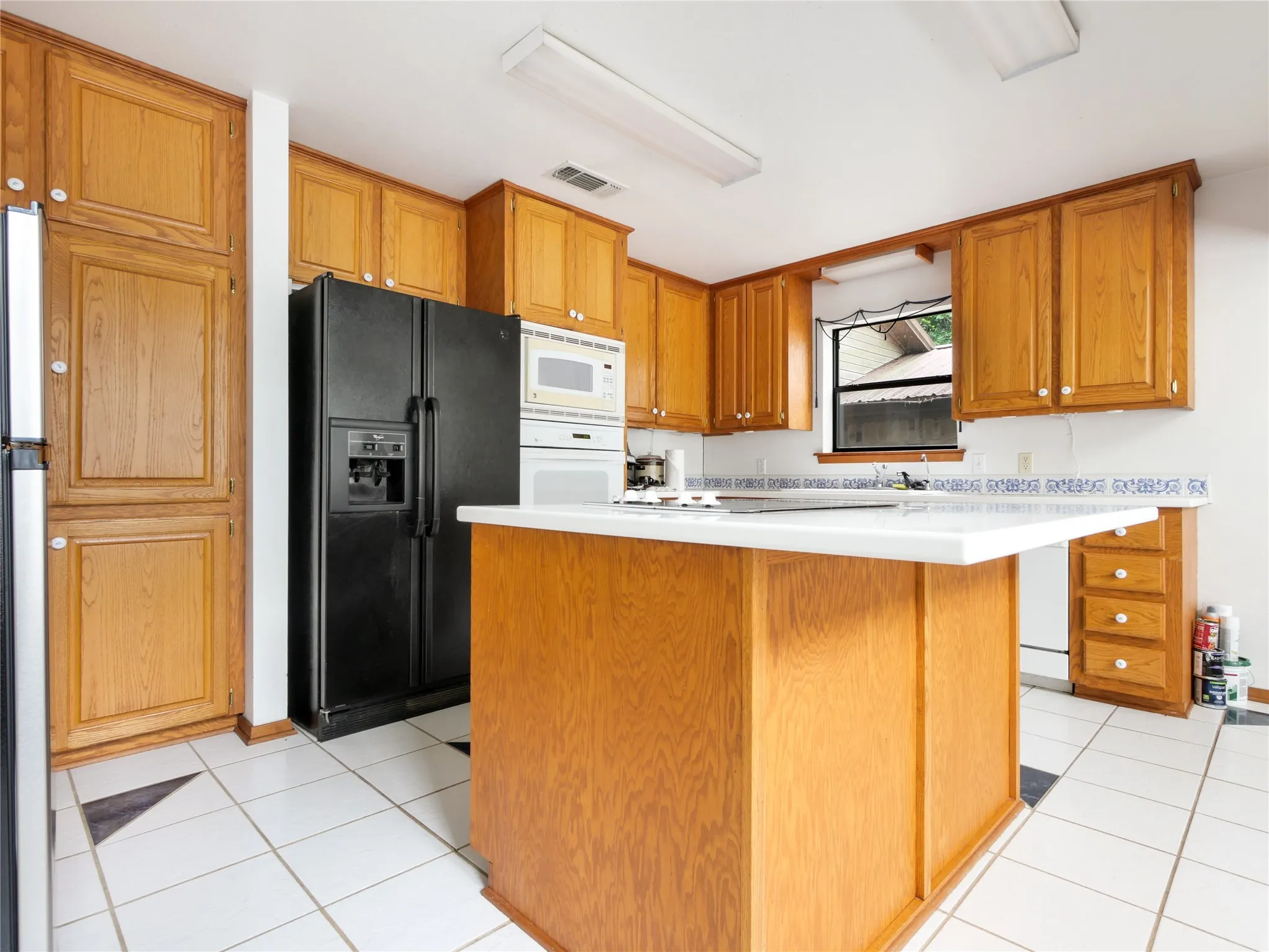 Kitchen featuring a central island with white countertop, extensive wood cabinetry, white tile flooring with dark accents, a built-in microwave, and a window above the sink