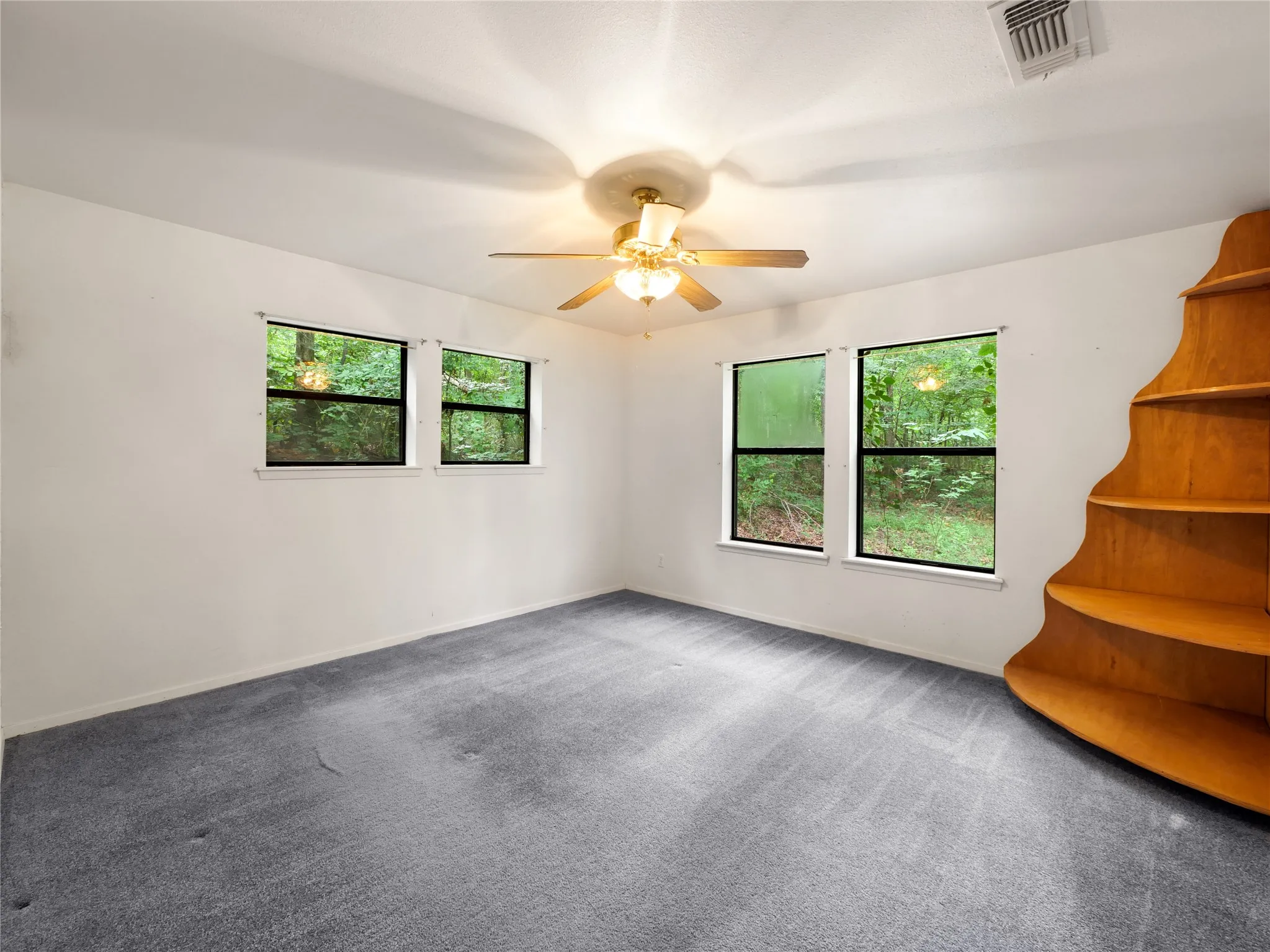 Spacious room featuring light gray carpeting, a built-in shelving unit with a curved design, multiple windows with dark frames, and a ceiling fan with integrated lighting