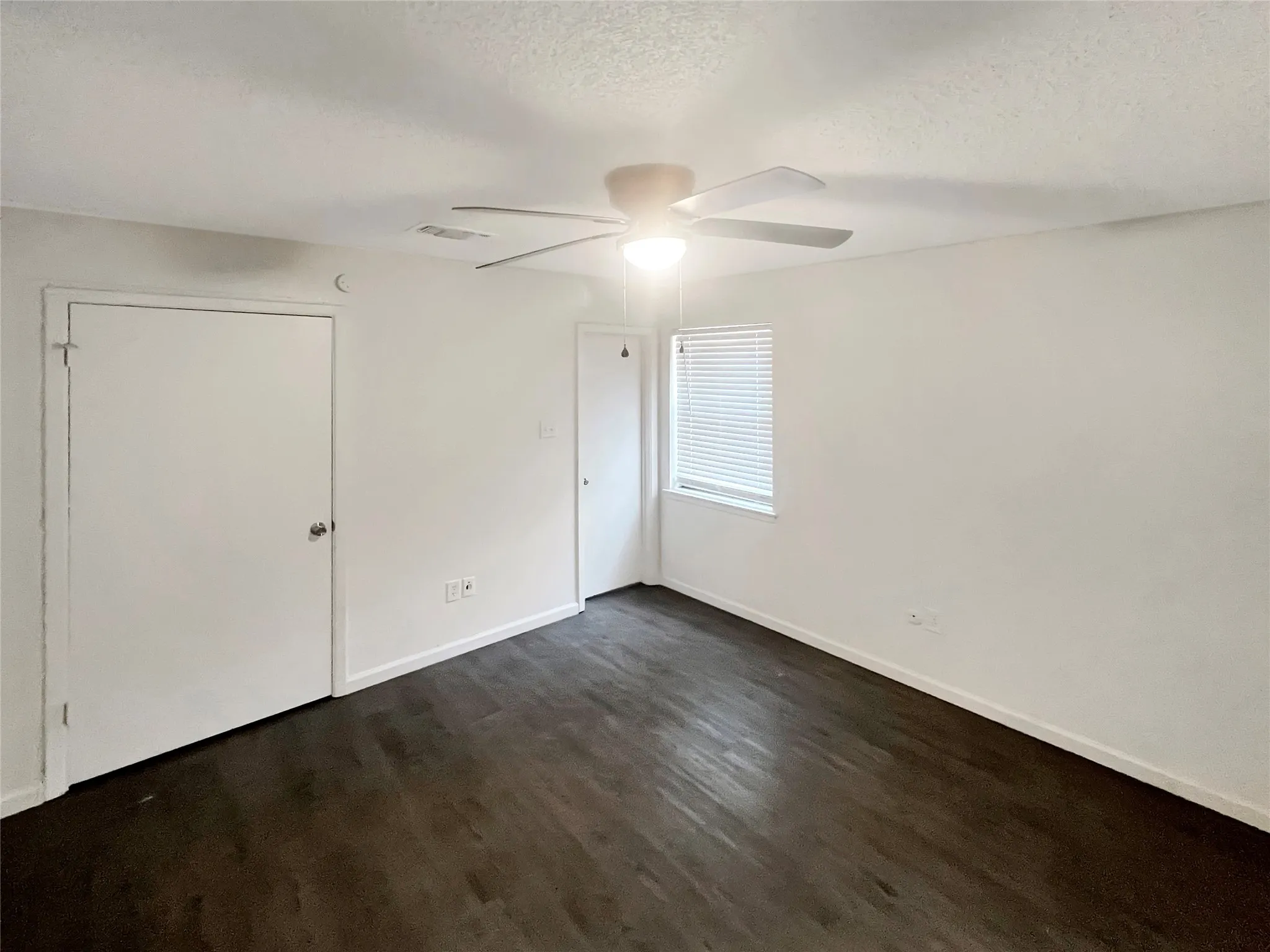 Spacious room featuring dark wood-finish flooring, white walls, and a ceiling fan with integrated lighting
