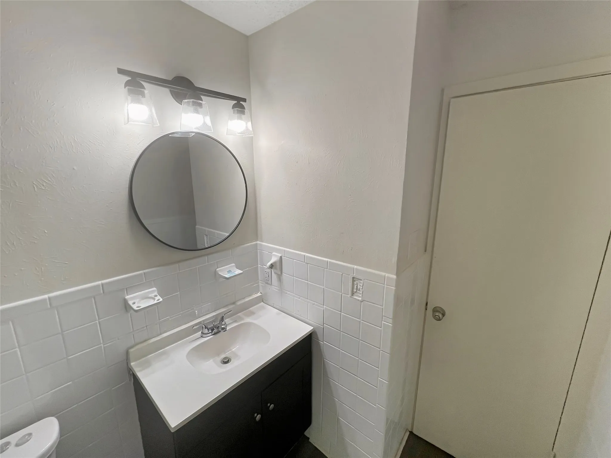 Bathroom featuring a circular mirror, matte black vanity light fixture, white subway tile wainscoting, a dark wood-finish vanity with an integrated sink, and a white countertop