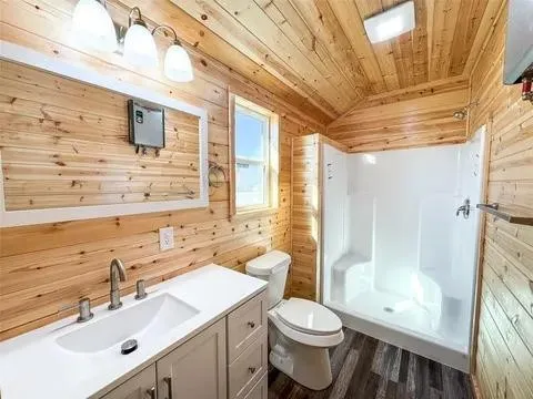 Bathroom featuring wood-finish walls and ceiling, a vanity with a rectangular undermount sink and two-handle faucet, a white toilet, and a white shower stall with built-in seating