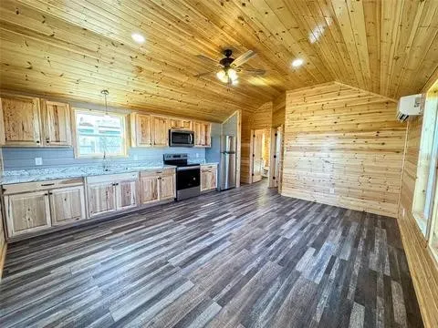 Open-concept living space with wood-finish flooring, natural wood plank walls and ceiling, and a full kitchen featuring light wood cabinetry, stone-look countertops, and stainless steel appliances including an oven range, microwave, and refrigerator