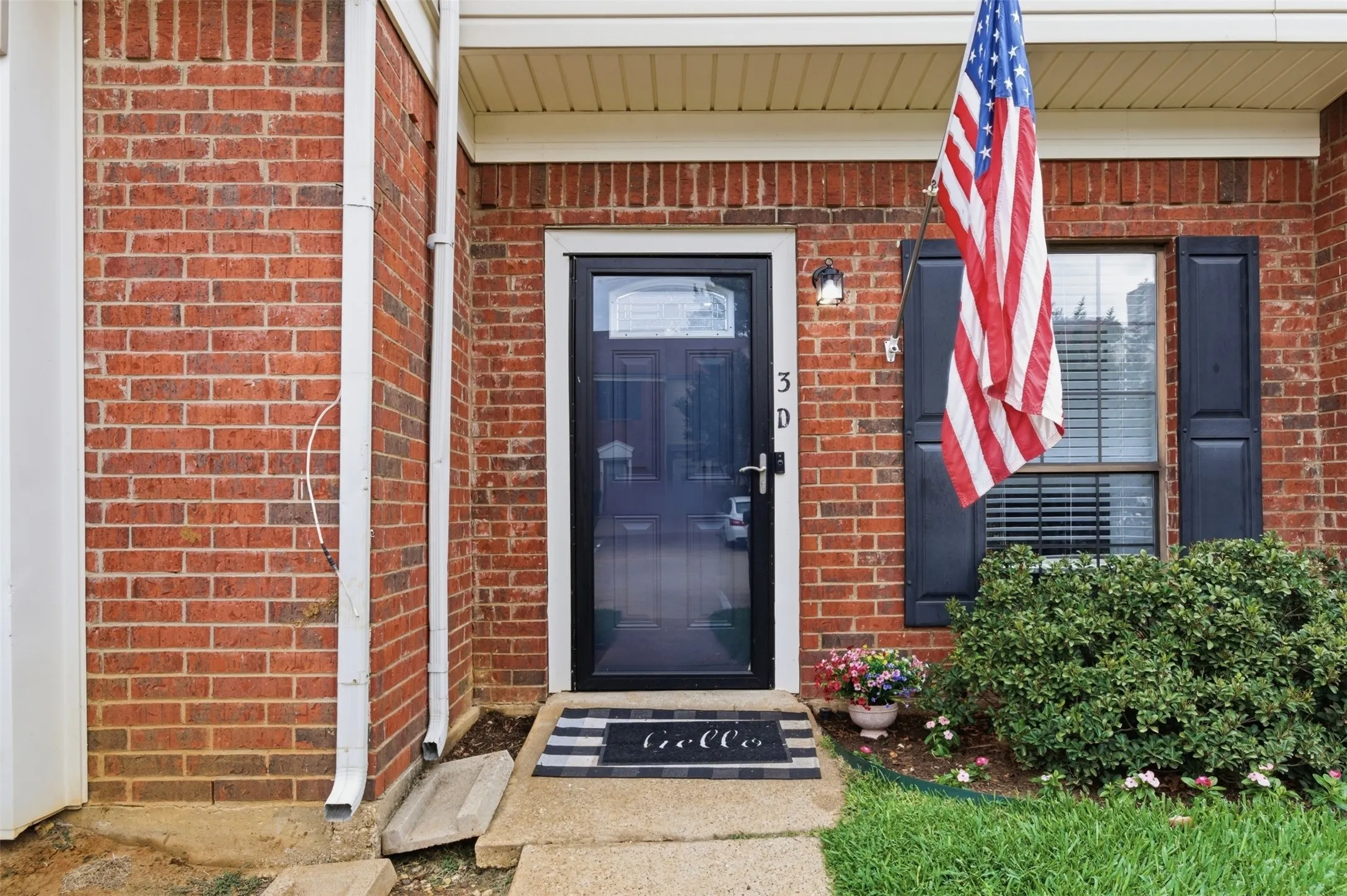 Brick exterior featuring a black front door with a glass panel, black window shutters, a front window with horizontal blinds, a concrete walkway, and established landscaping