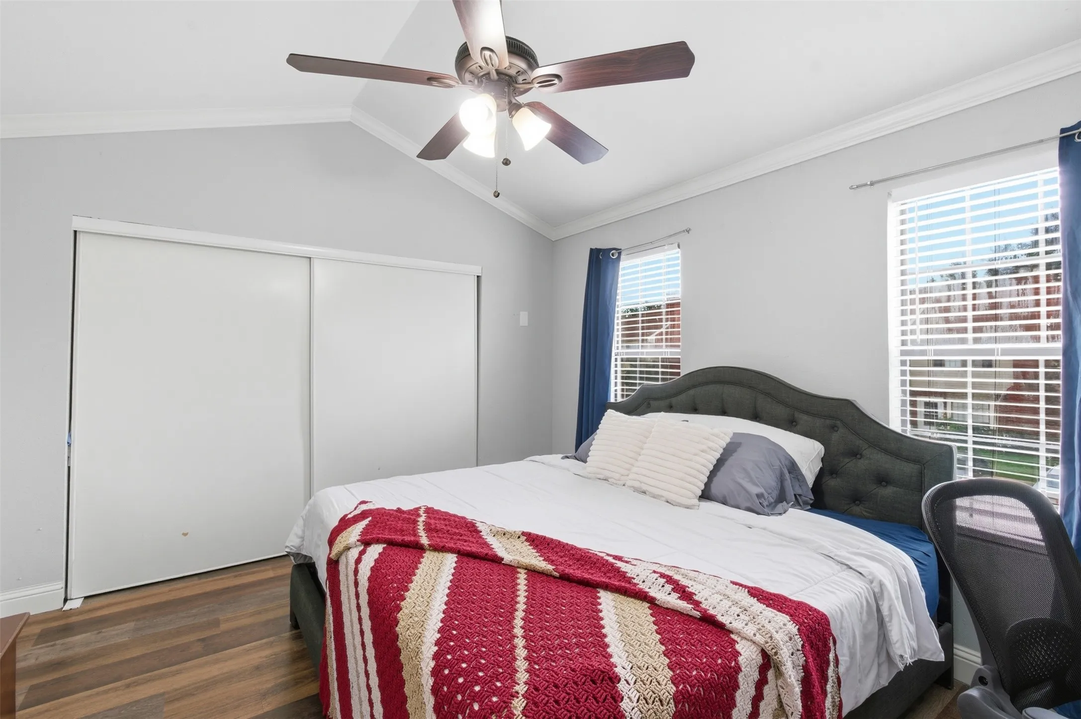 Vaulted ceiling bedroom with a ceiling fan light fixture, two windows with blinds, and wood-finish flooring