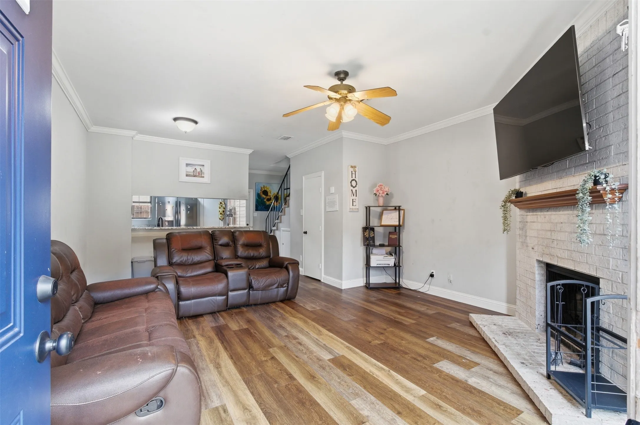 Living area featuring wood-finish flooring, a white brick fireplace with a wood mantel, crown molding, a ceiling fan, and a pass-through opening