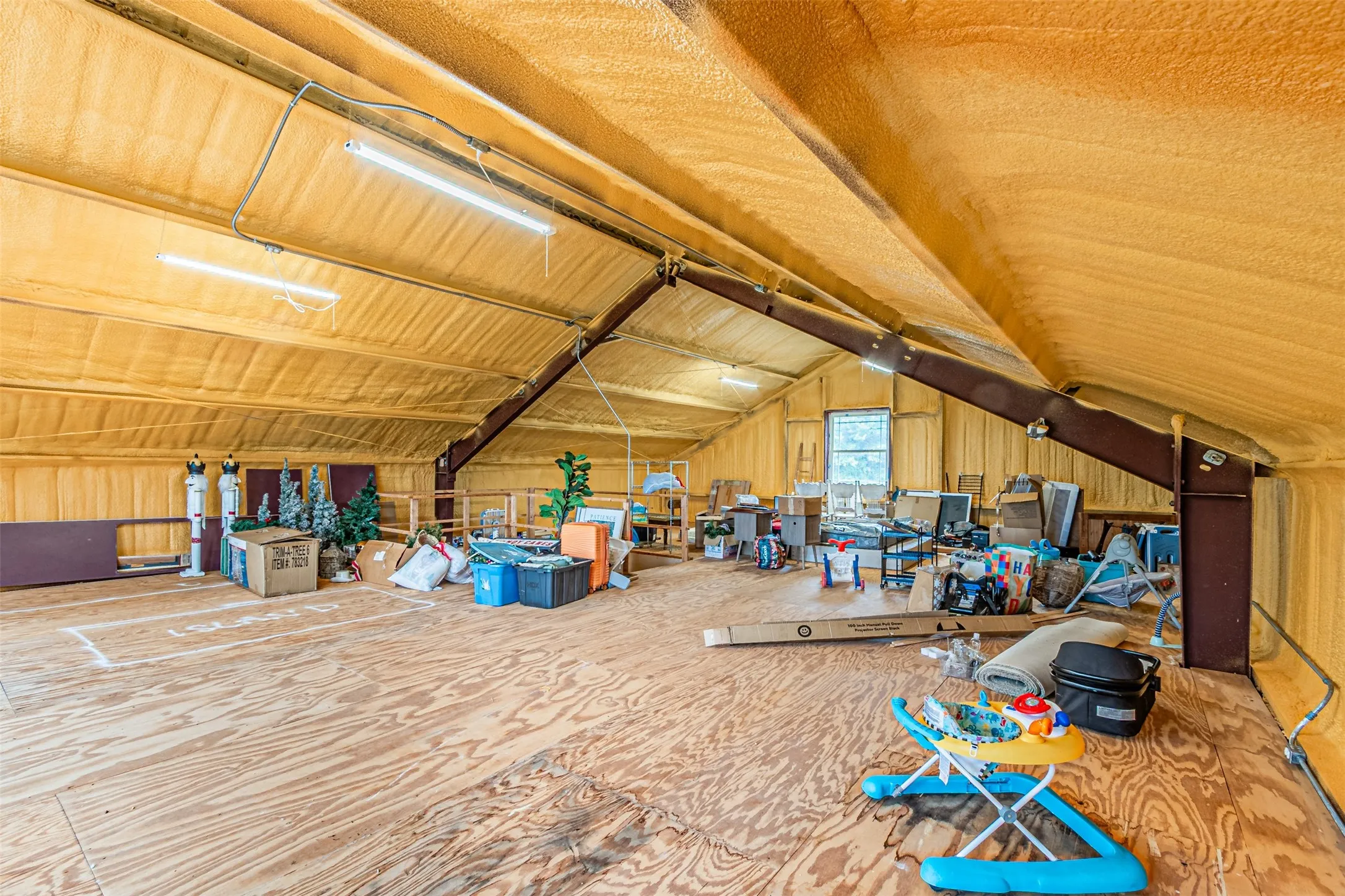 Spacious attic area featuring wood-finish subflooring, exposed structural beams, spray foam insulation, and a window providing natural light