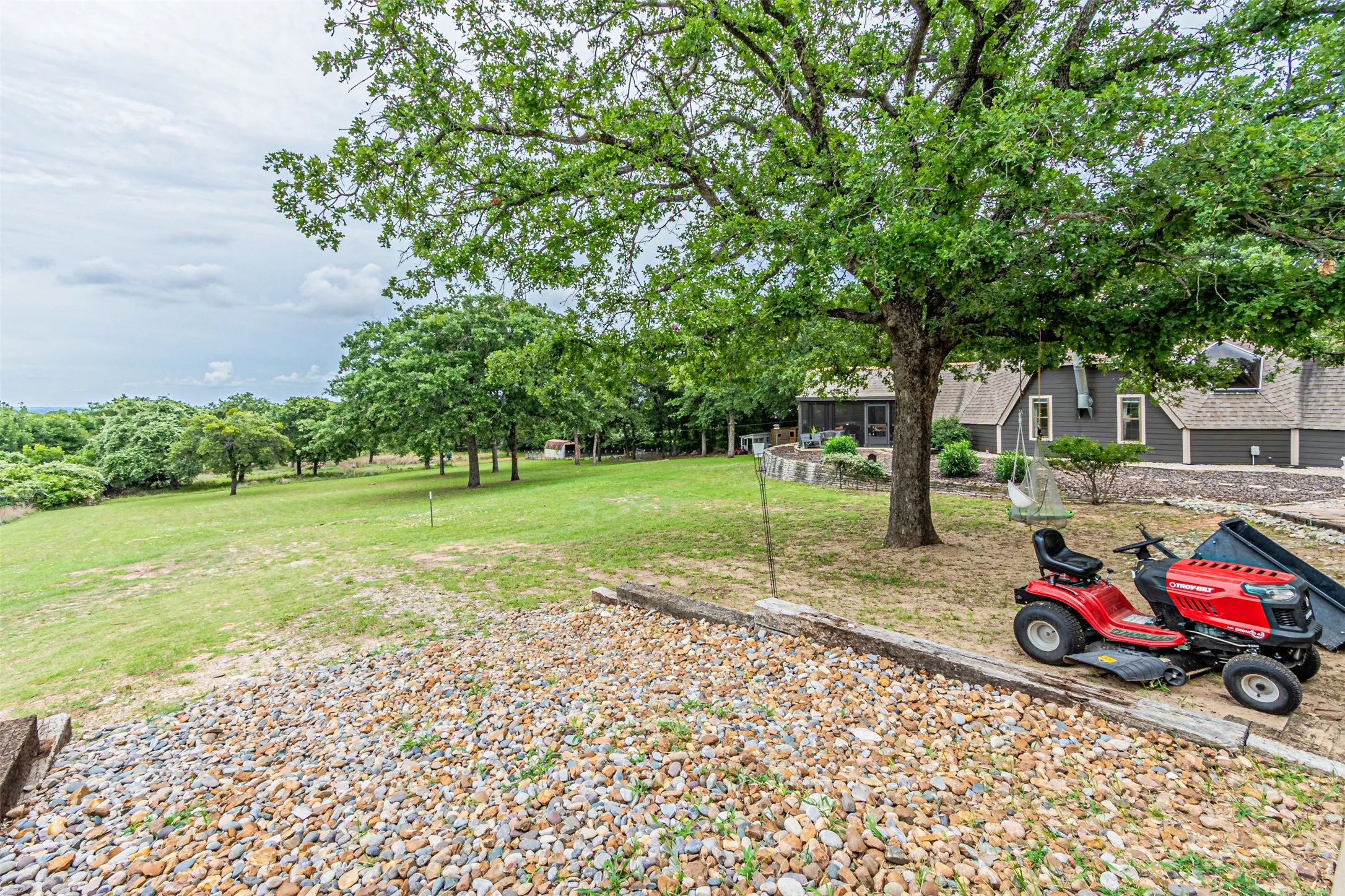 Expansive rear yard featuring a gravel retention area, mature trees, and a sloped lawn extending into a wooded area