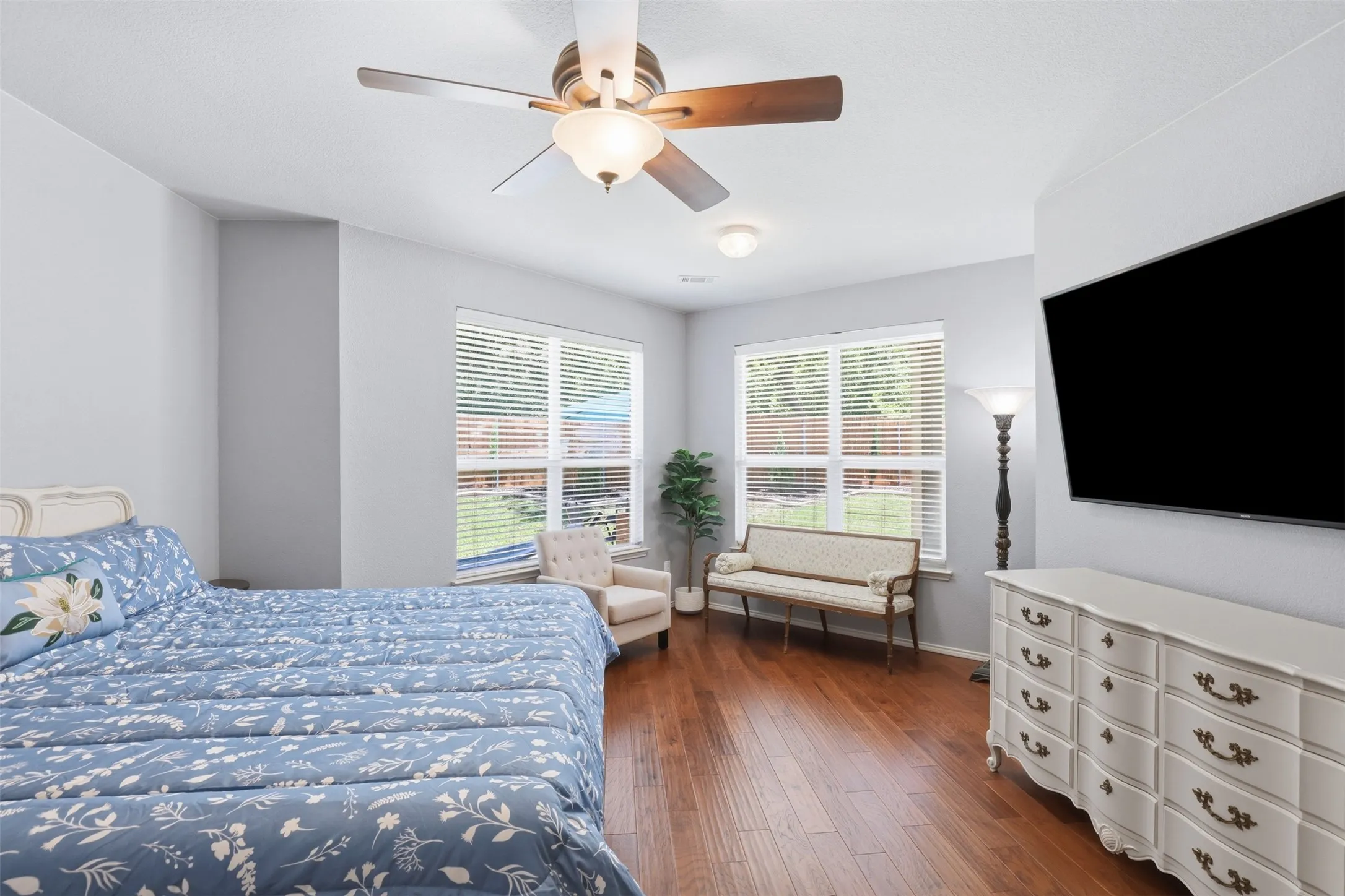 Primary bedroom featuring wood-finish flooring, two large windows with horizontal blinds, and a ceiling fan with integrated lighting