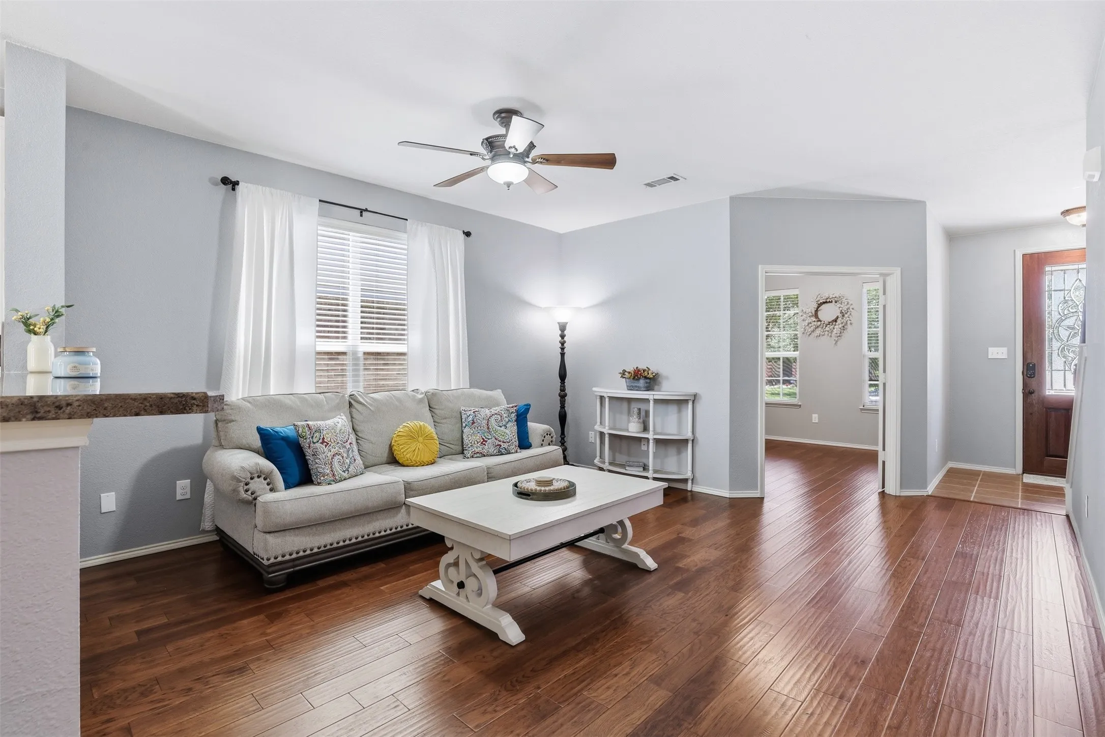 Living area featuring rich wood-finish flooring, light grey walls, and a ceiling fan with integrated lighting
