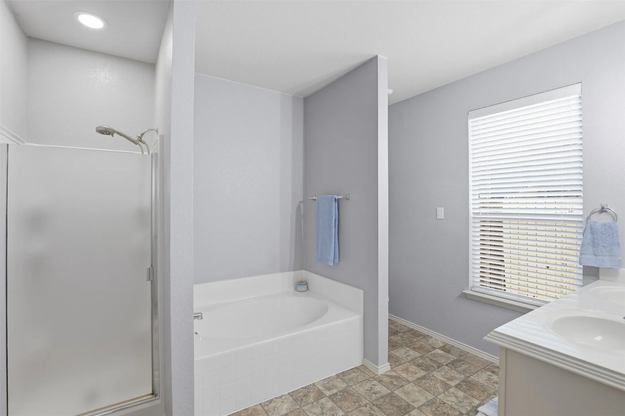 Primary bathroom featuring a built-in soaking tub, a shower enclosure with frosted glass, a window with horizontal blinds, and a vanity with dual sinks