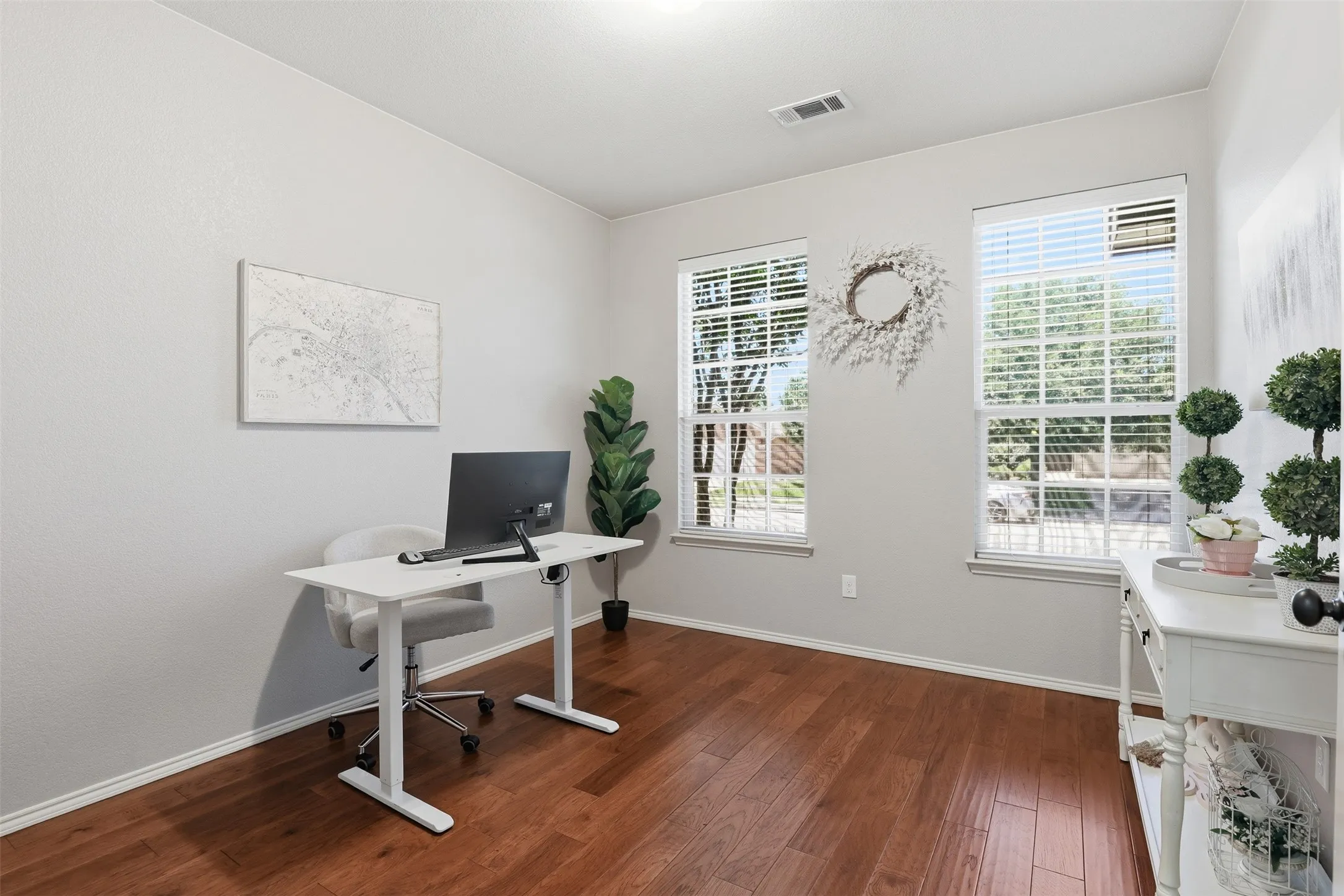 Hardwood-finish flooring complements light gray walls in this versatile room featuring two large windows with blinds