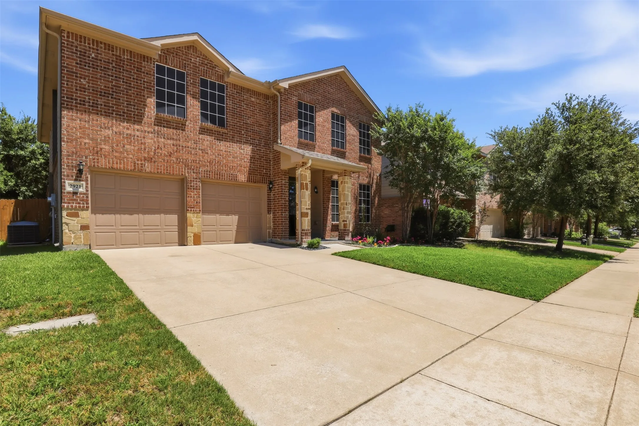 Brick exterior with stone accents, featuring a two-car garage, mature front yard trees, a concrete driveway, and a sidewalk