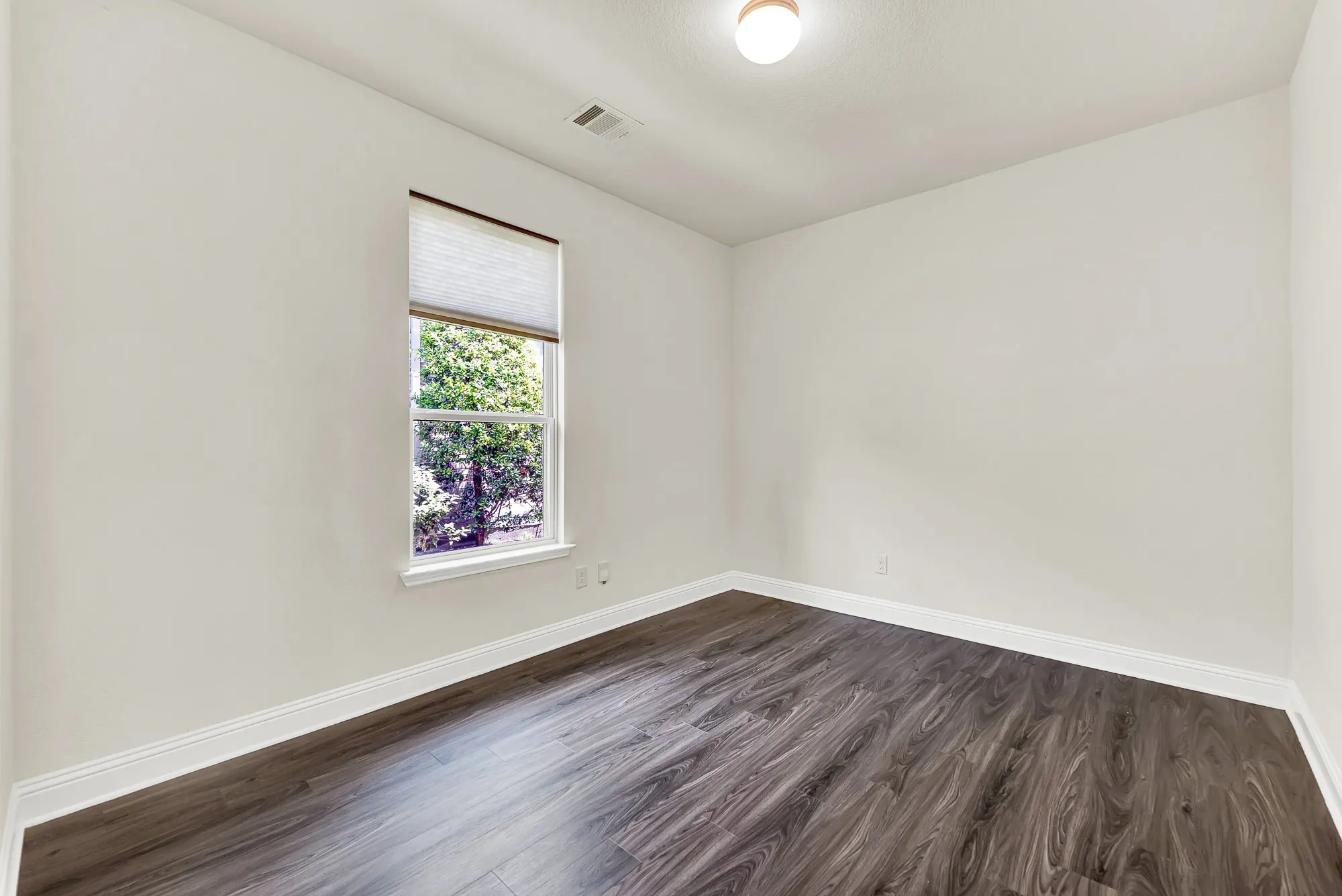 A single, well-placed window frames a view of green foliage, instantly softening the clean lines of the cool-toned plank flooring. This versatile space is ideal for setting up a quiet afternoon yoga session or an inspiring, distraction-free home office setup