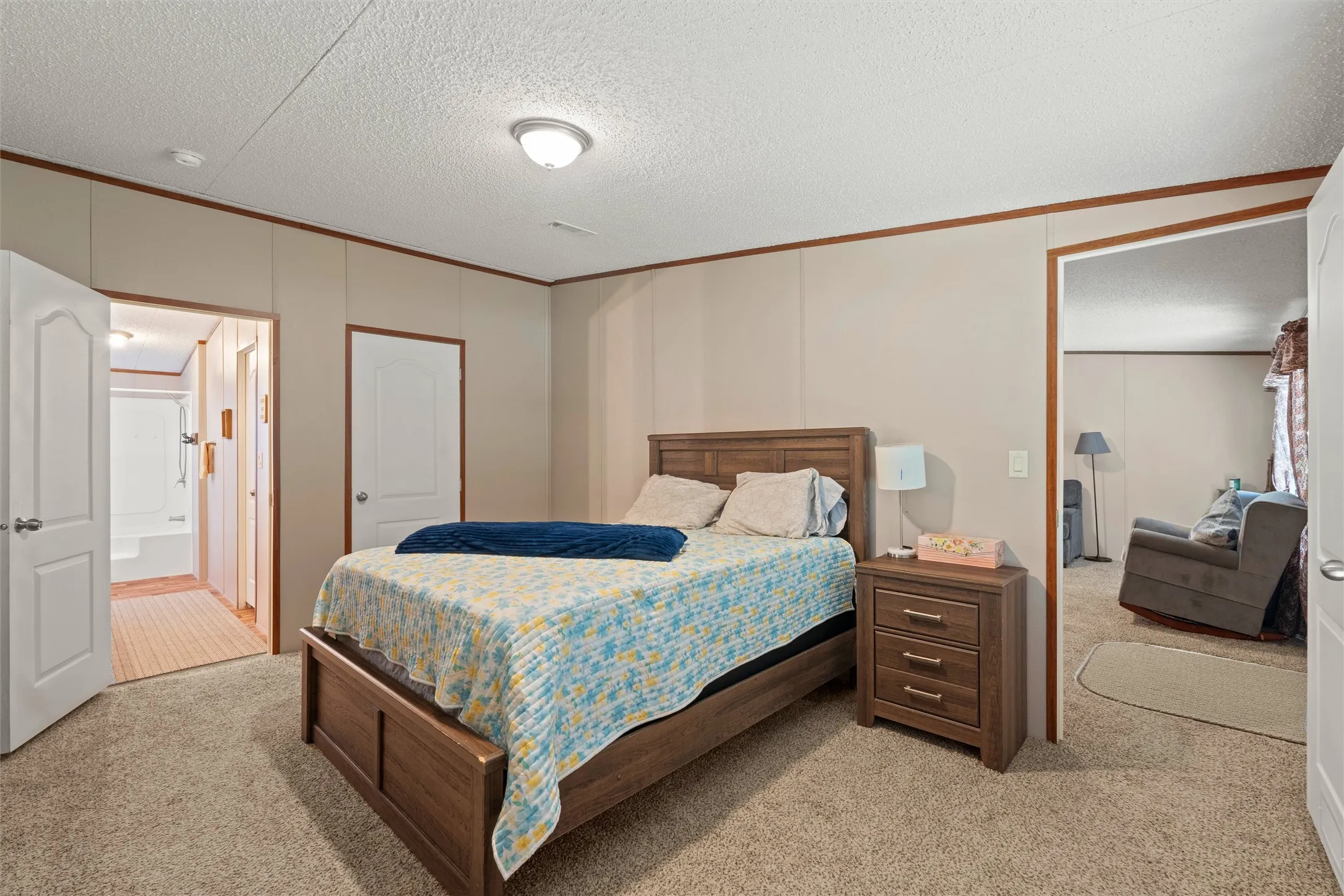 Carpeted bedroom featuring neutral wall panels with wood-finish trim, a flush-mount ceiling light fixture, and an open doorway revealing an adjacent room