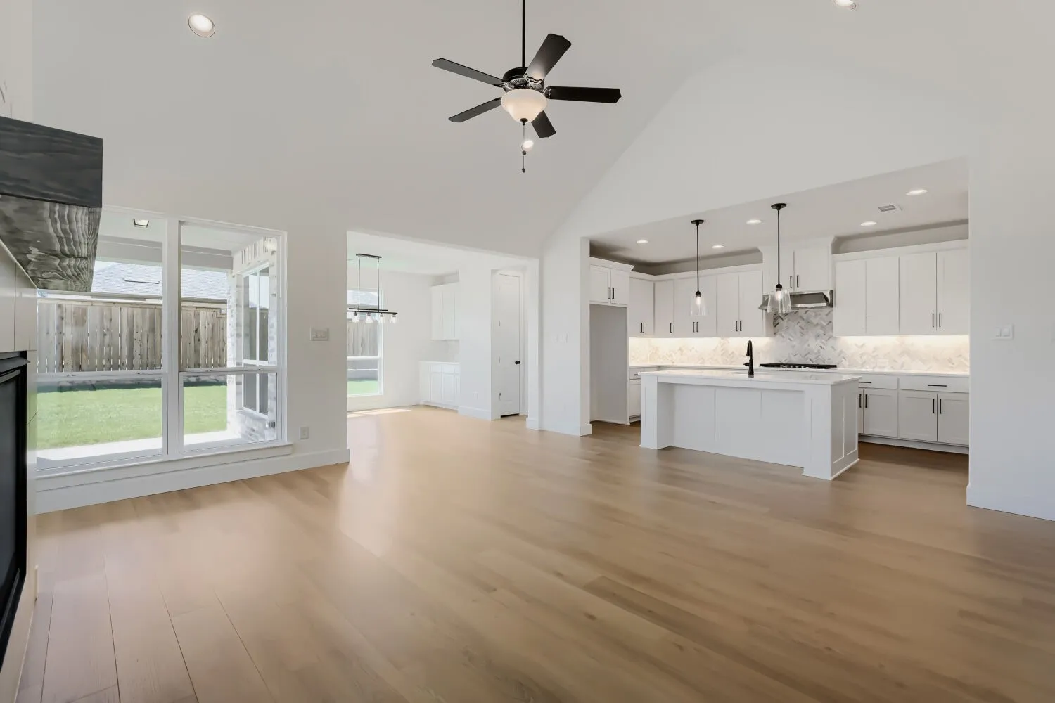 Vaulted ceiling with a ceiling fan, light wood-finish flooring, white kitchen cabinetry with pendant lighting, kitchen island with a built-in sink, and a large window overlooking a manicured lawn