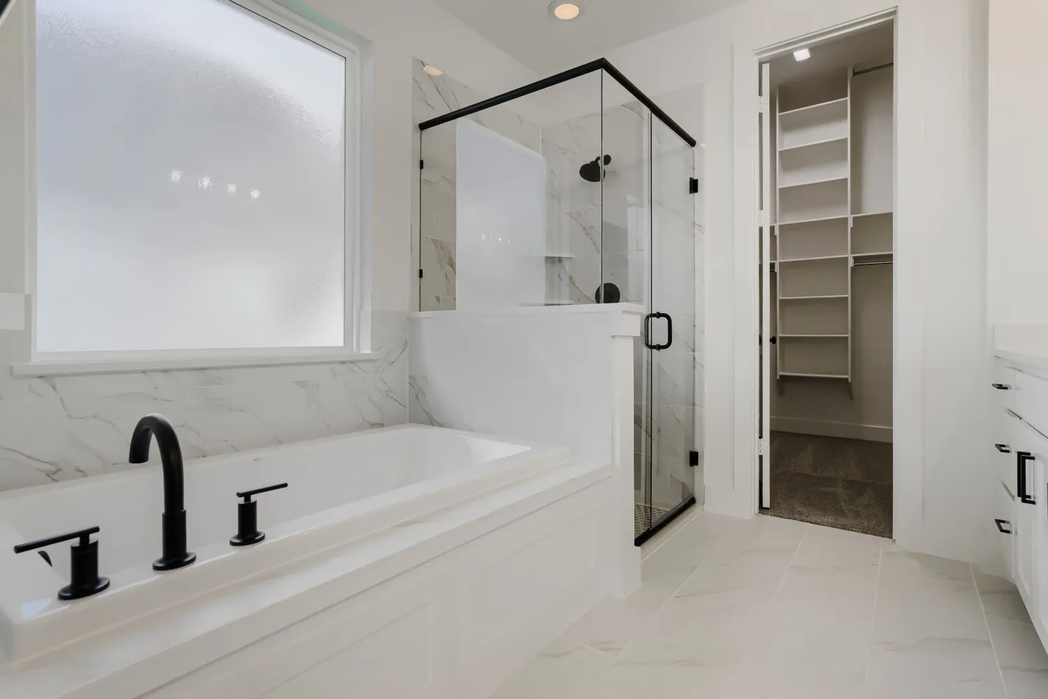 Soaking tub with matte black fixtures, glass-enclosed shower, white tile flooring, and a walk-in closet with built-in shelving