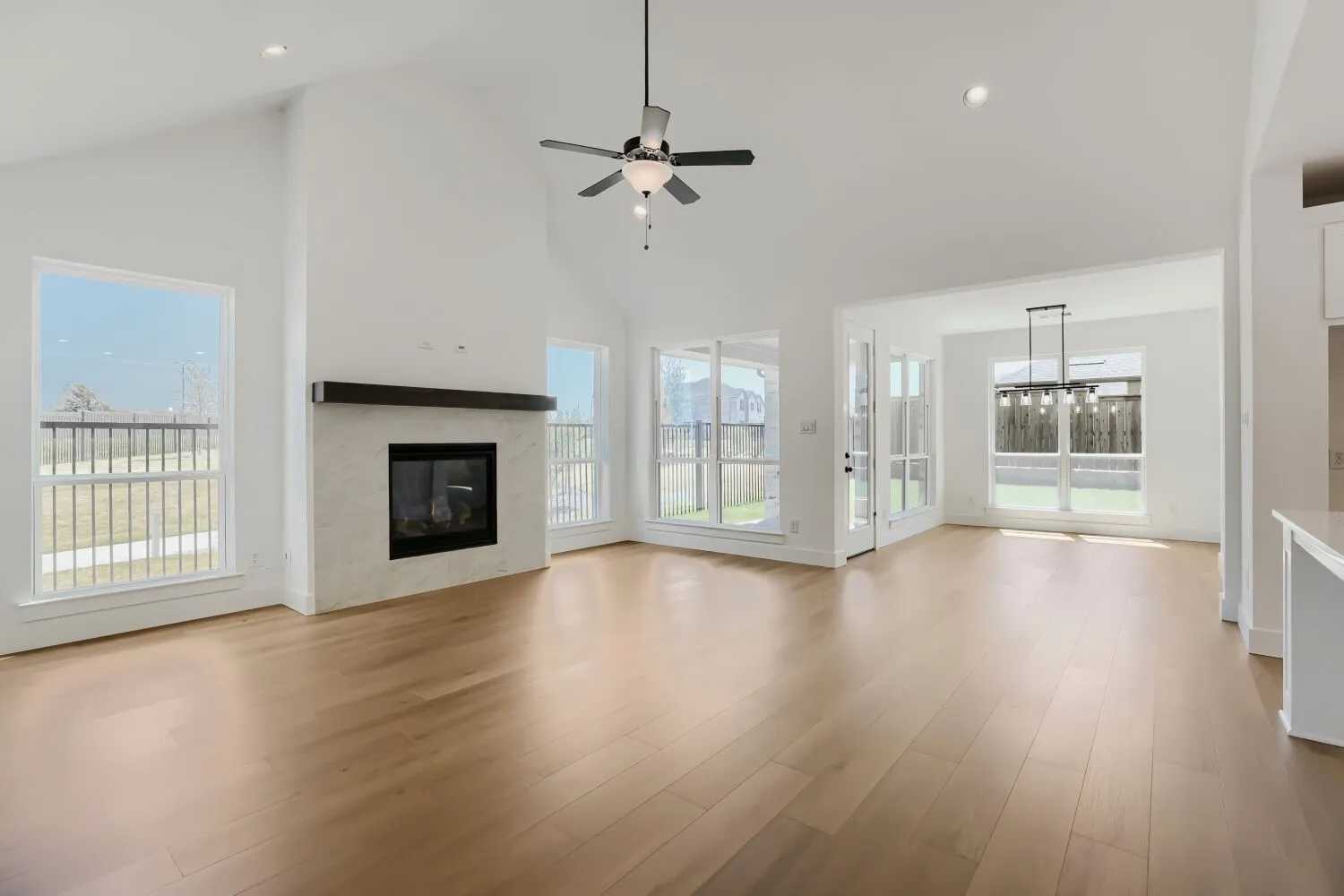 Vaulted ceiling with a ceiling fan, fireplace with a stone surround and wood mantel, light wood-finish flooring, multiple large windows, and a glass-paneled door