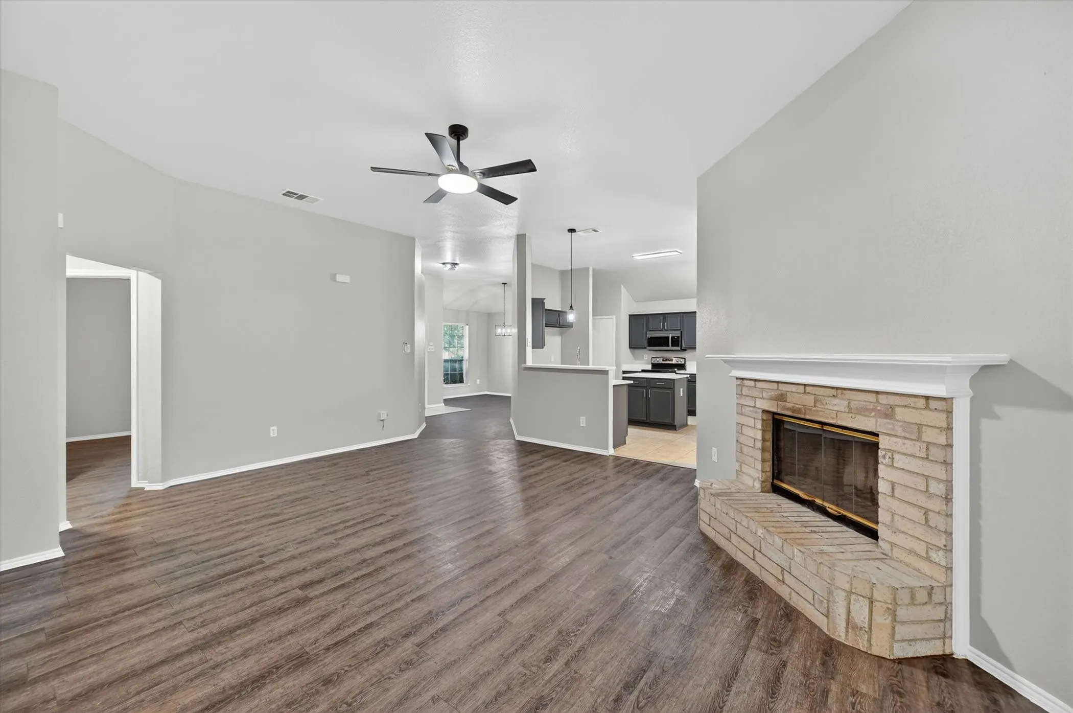 Spacious living area featuring a corner brick fireplace with a white mantel, wood-finish flooring, and a ceiling fan