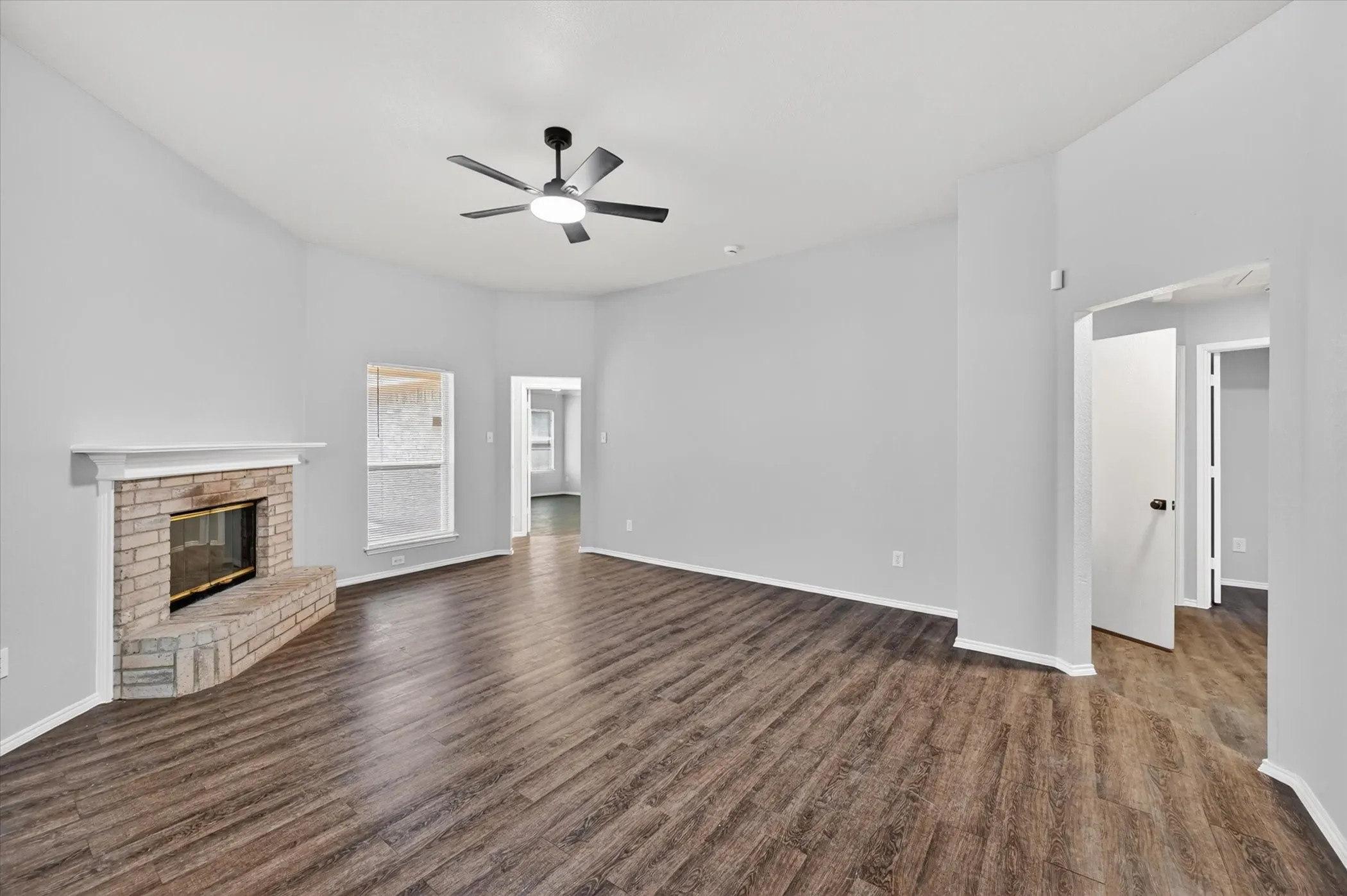 Spacious living area featuring wood-finish flooring, a brick-surround fireplace with a white mantel, and a ceiling fan with integrated lighting