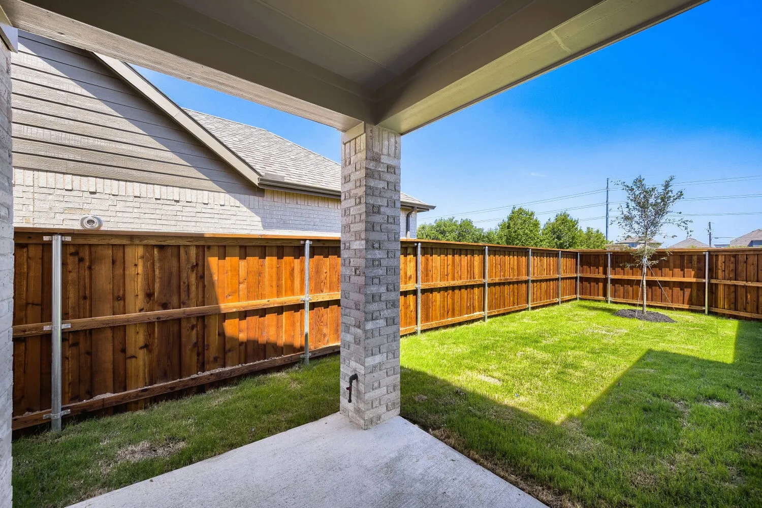 Covered patio with a concrete surface, overlooking a fenced backyard featuring a young tree and a manicured lawn