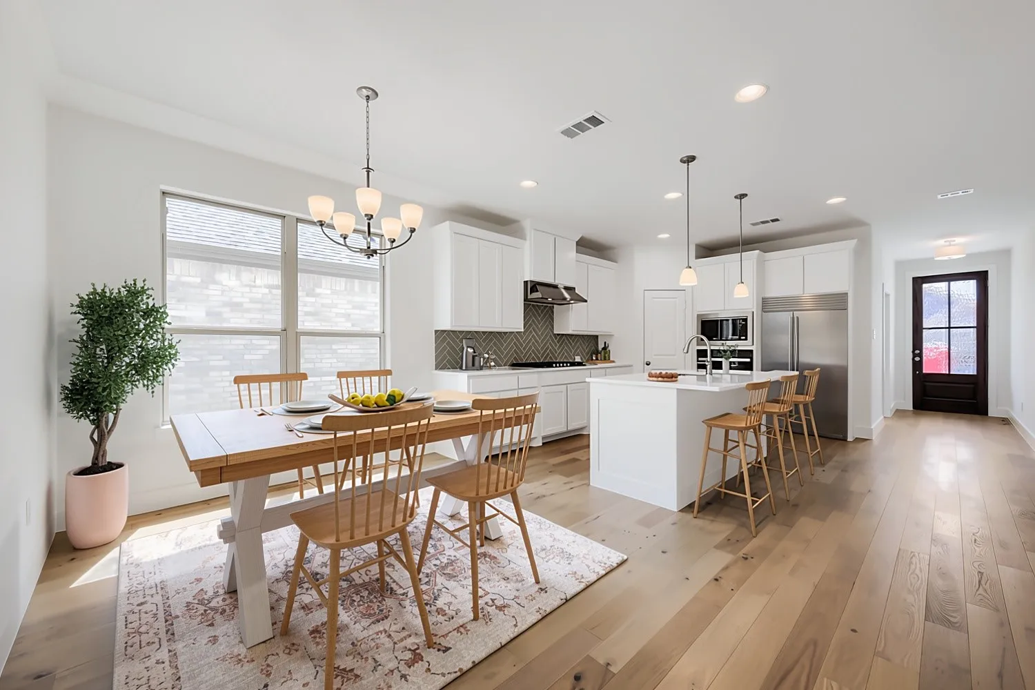 Virtually Staged Photo - Open-concept kitchen and dining area featuring white cabinetry, stainless steel appliances, waterfall-edge island with seating, wide-plank wood-finish flooring, large windows with natural light, and a coordinating chandelier fixture