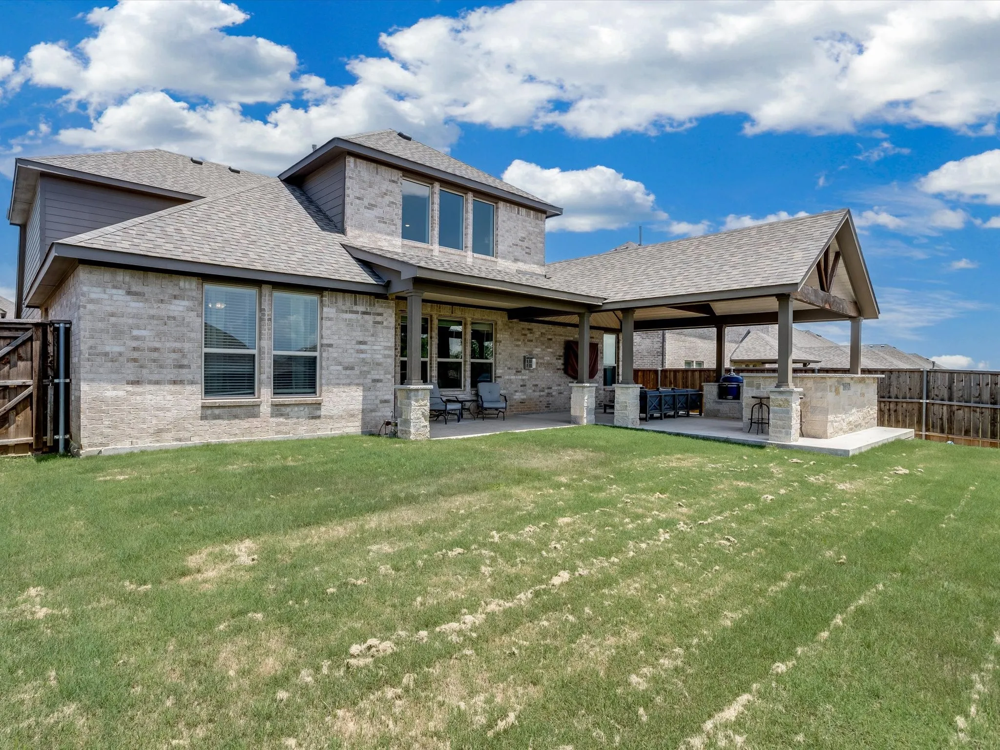 Rear exterior featuring a covered patio, outdoor kitchen, stone accent columns, brick and siding facade, and a fenced perimeter