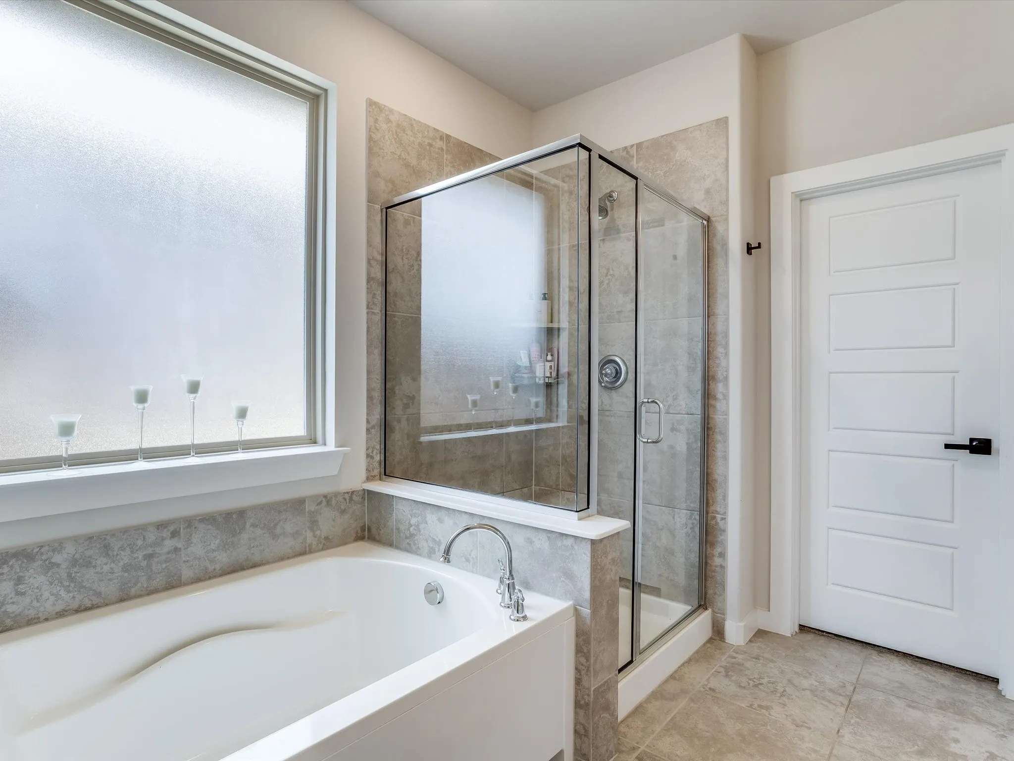 Bathroom featuring a built-in soaking tub with chrome fixtures, a clear glass enclosed shower with a built-in niche, light-toned tile flooring, and a modern white paneled door with matte black hardware