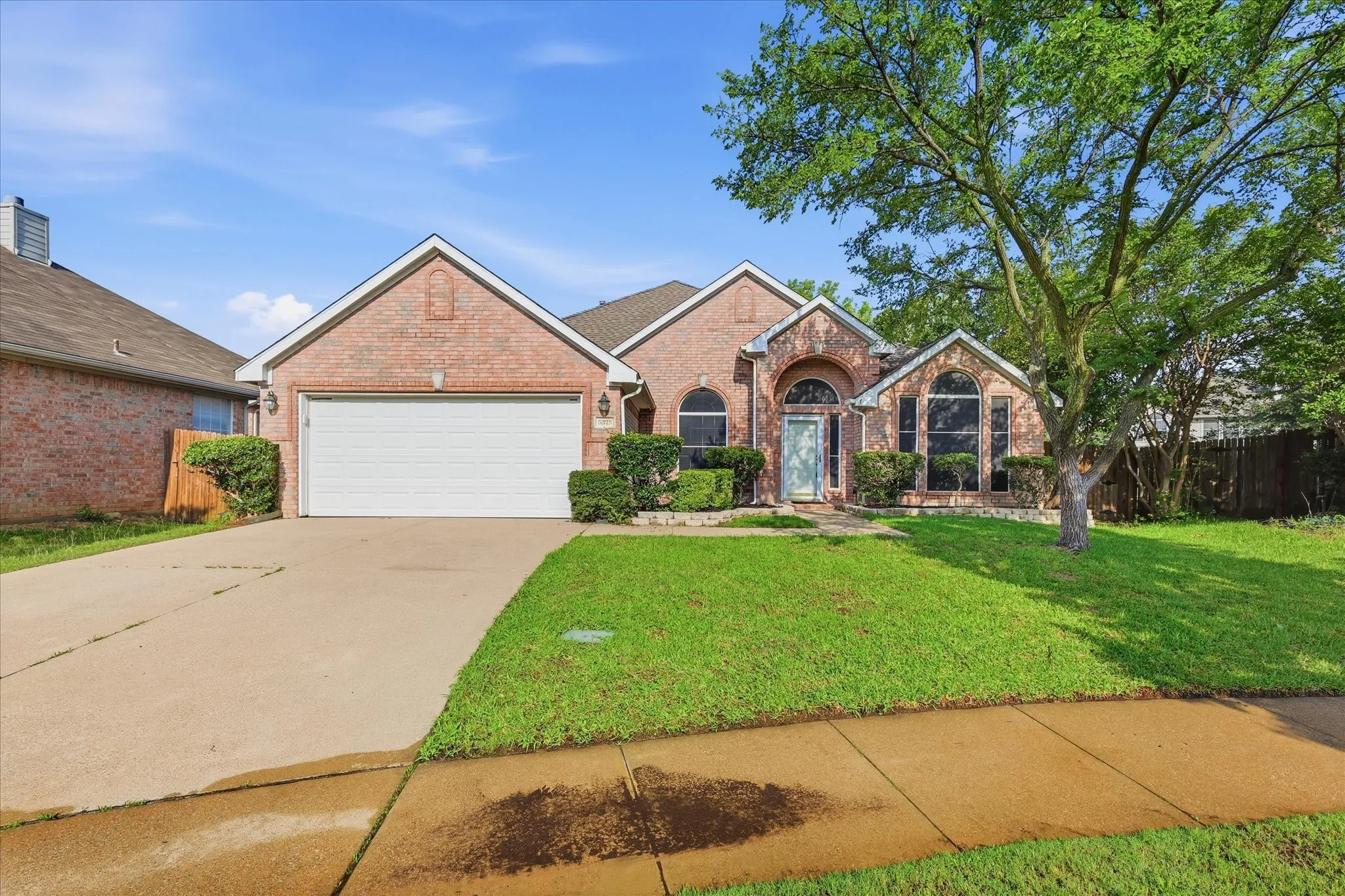 Brick exterior residence featuring an arched entry, a two-car garage, established front landscaping, an expansive driveway, and a mature shade tree