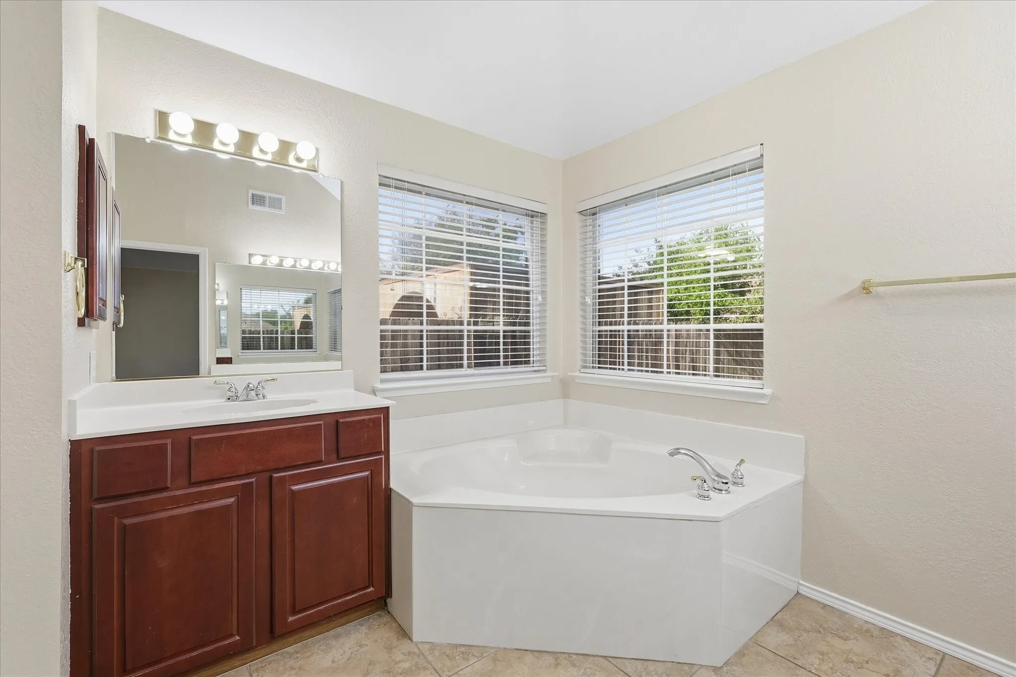 Bathroom featuring a corner soaking tub with deck-mounted fixtures, two windows with blinds, a single vanity with a white countertop and dark wood-finish cabinetry, and a large wall mirror with overhead vanity lighting