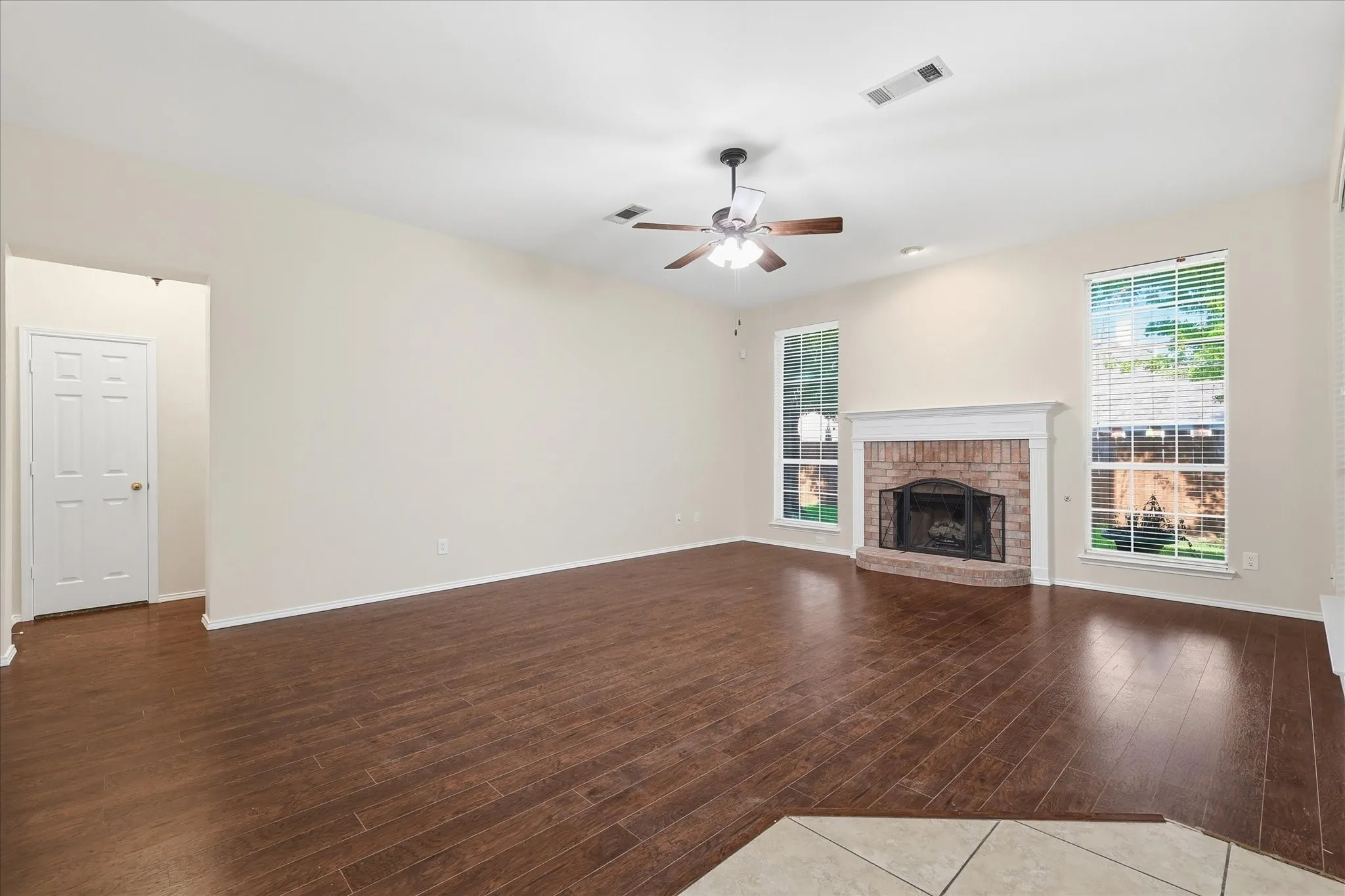 Spacious living area featuring rich wood-finish flooring, a brick fireplace with a white mantel, and multiple windows providing natural illumination