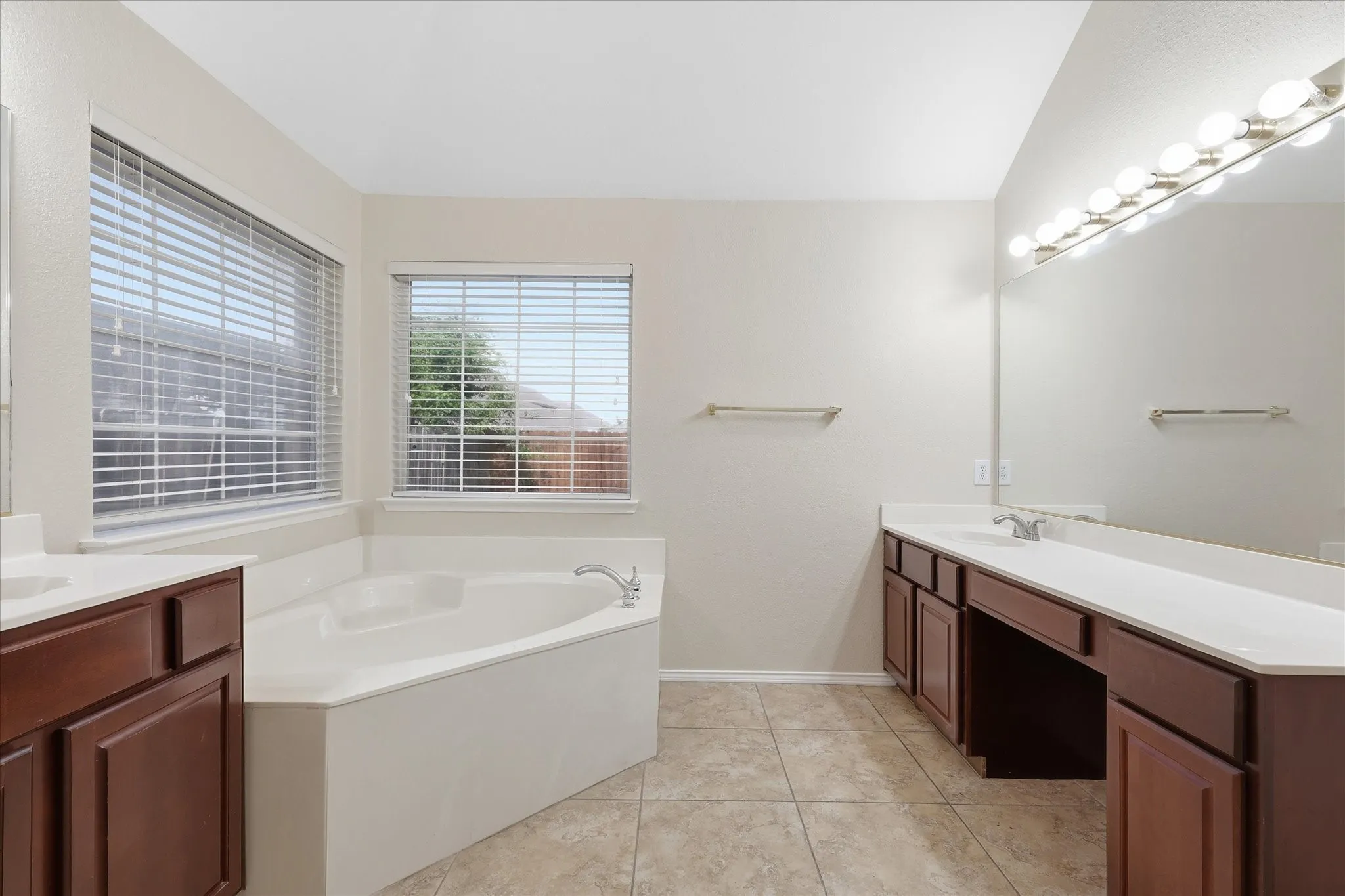 Corner soaking tub with dual windows, complemented by a dual vanity featuring dark wood cabinetry, white countertops, and tile flooring
