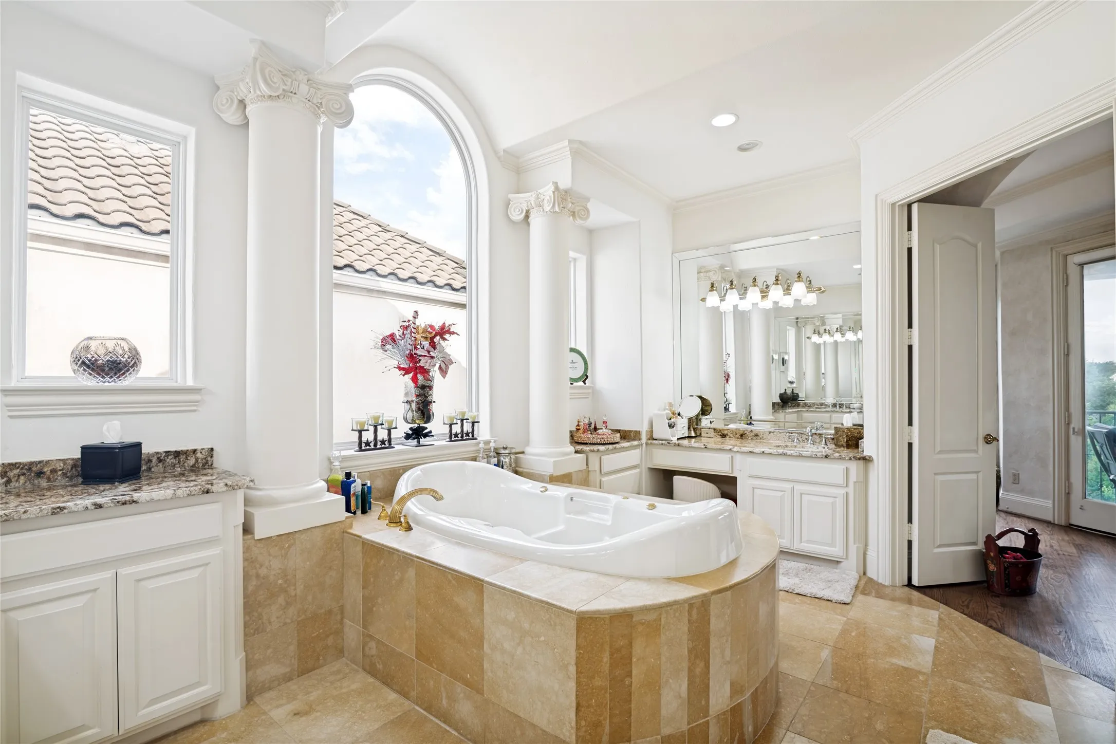 Spa-inspired bathroom featuring a built-in soaking tub, arched window, column architectural details, dual vanity with undermount sinks, and stone tile flooring