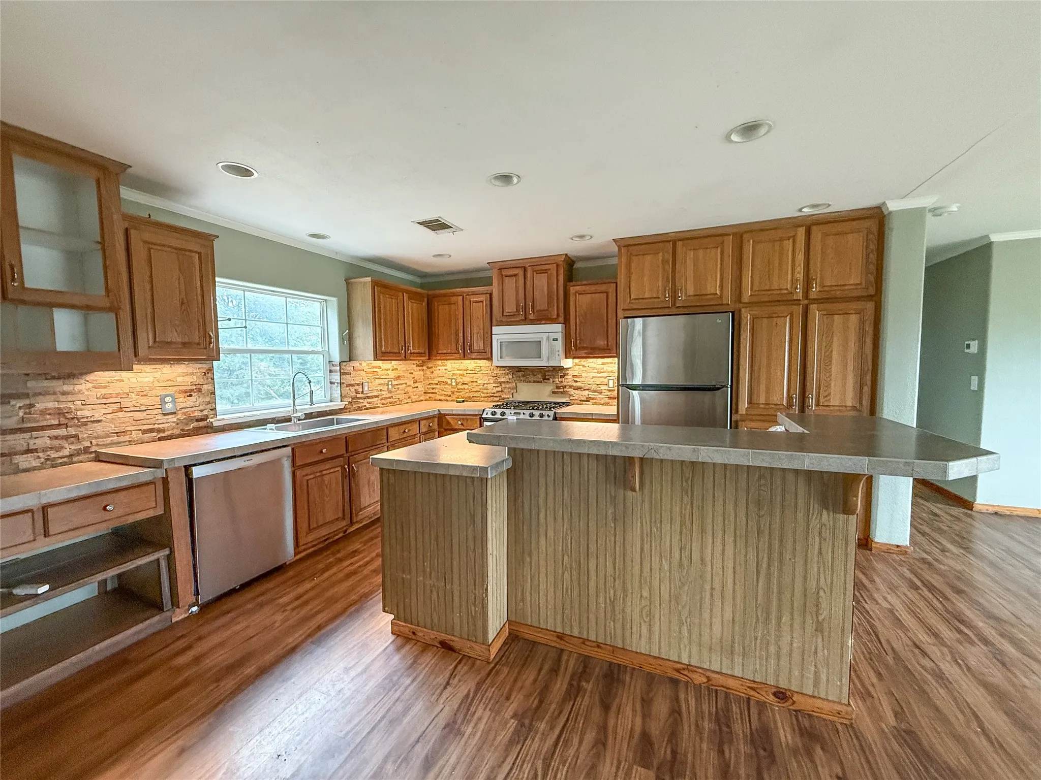 Kitchen featuring wood-finish cabinetry, stainless steel appliances, a stone tile backsplash, recessed lighting, and an island with a breakfast bar
