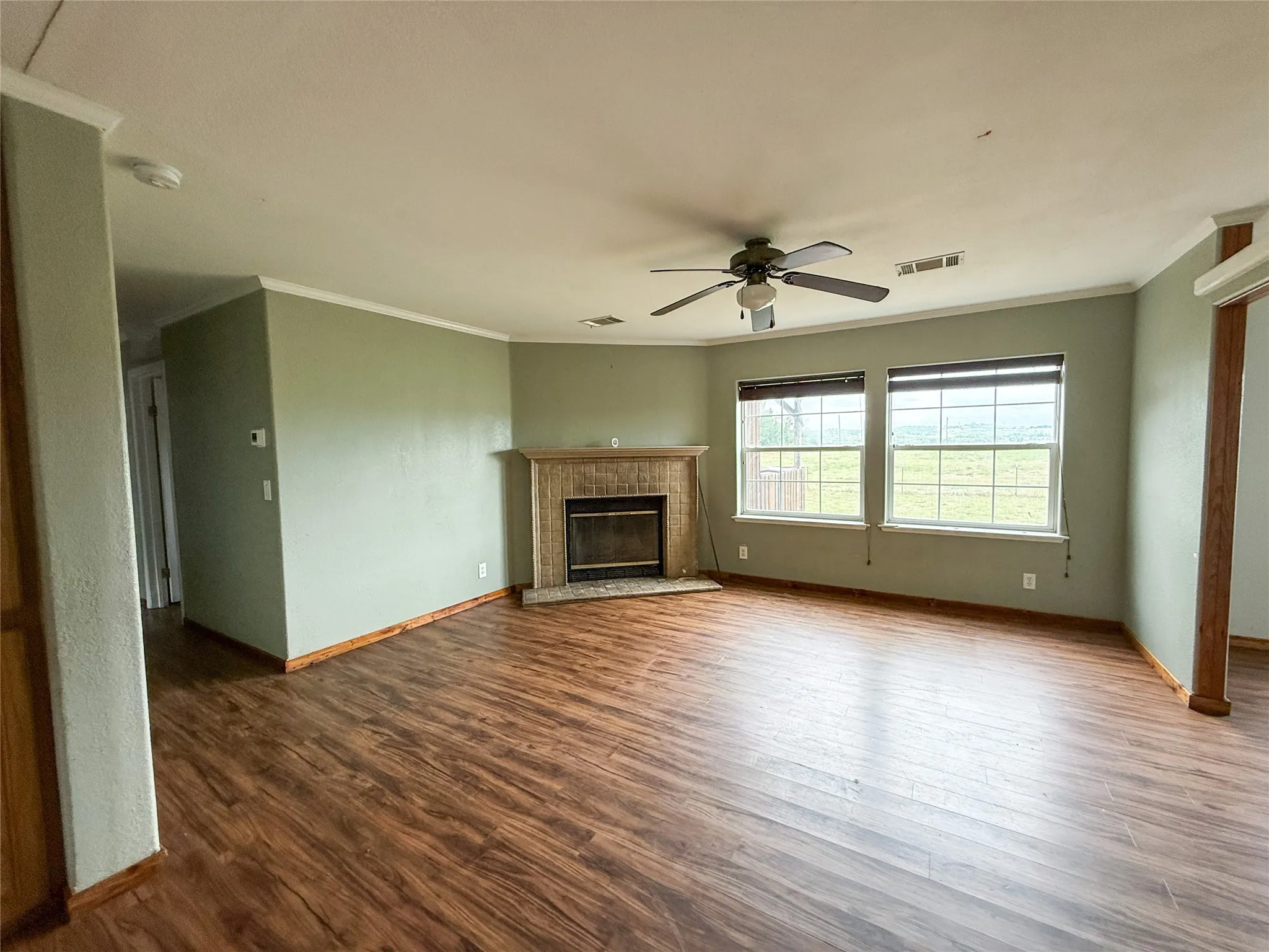 Spacious living area featuring wood-finish flooring, a fireplace with a tiled surround and wood mantel, two large windows, a ceiling fan, and crown molding