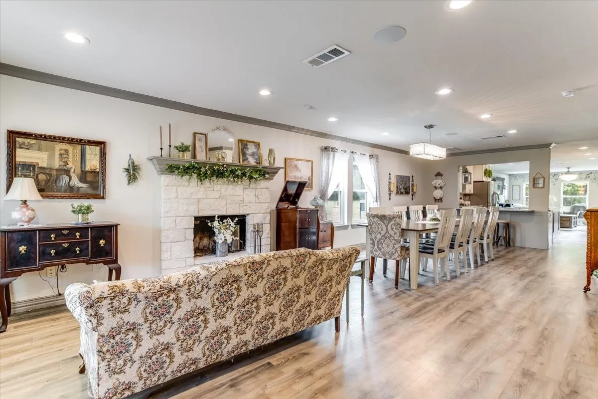 Expansive open-concept living area featuring light wood-finish flooring, a stone-faced fireplace with a mantel, recessed lighting, and a modern dining chandelier