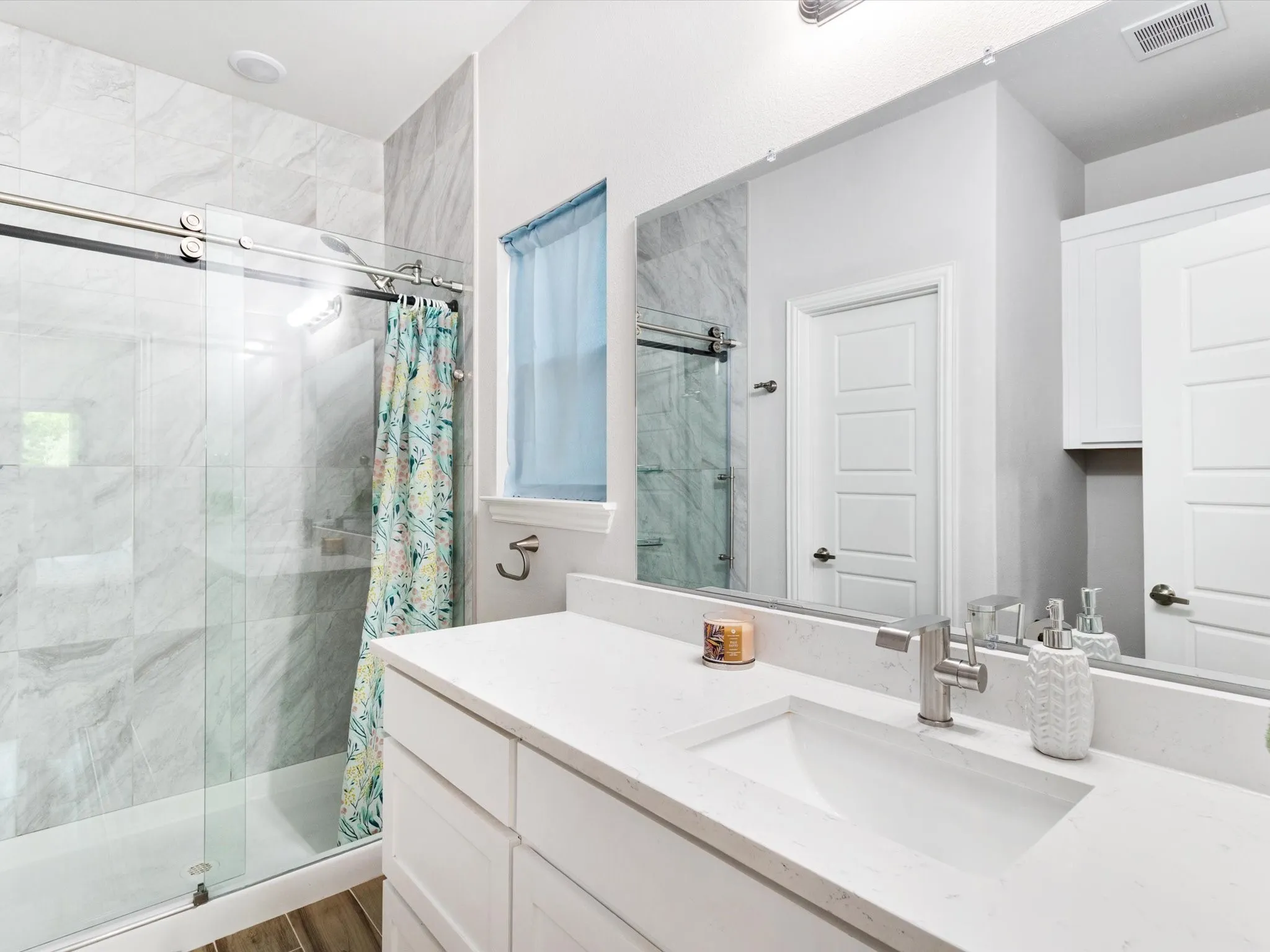 Bathroom featuring a spacious vanity with an undermount sink, a large mirror, white cabinetry, a window with frosted glass, and a tub with a sliding glass door and tile surround