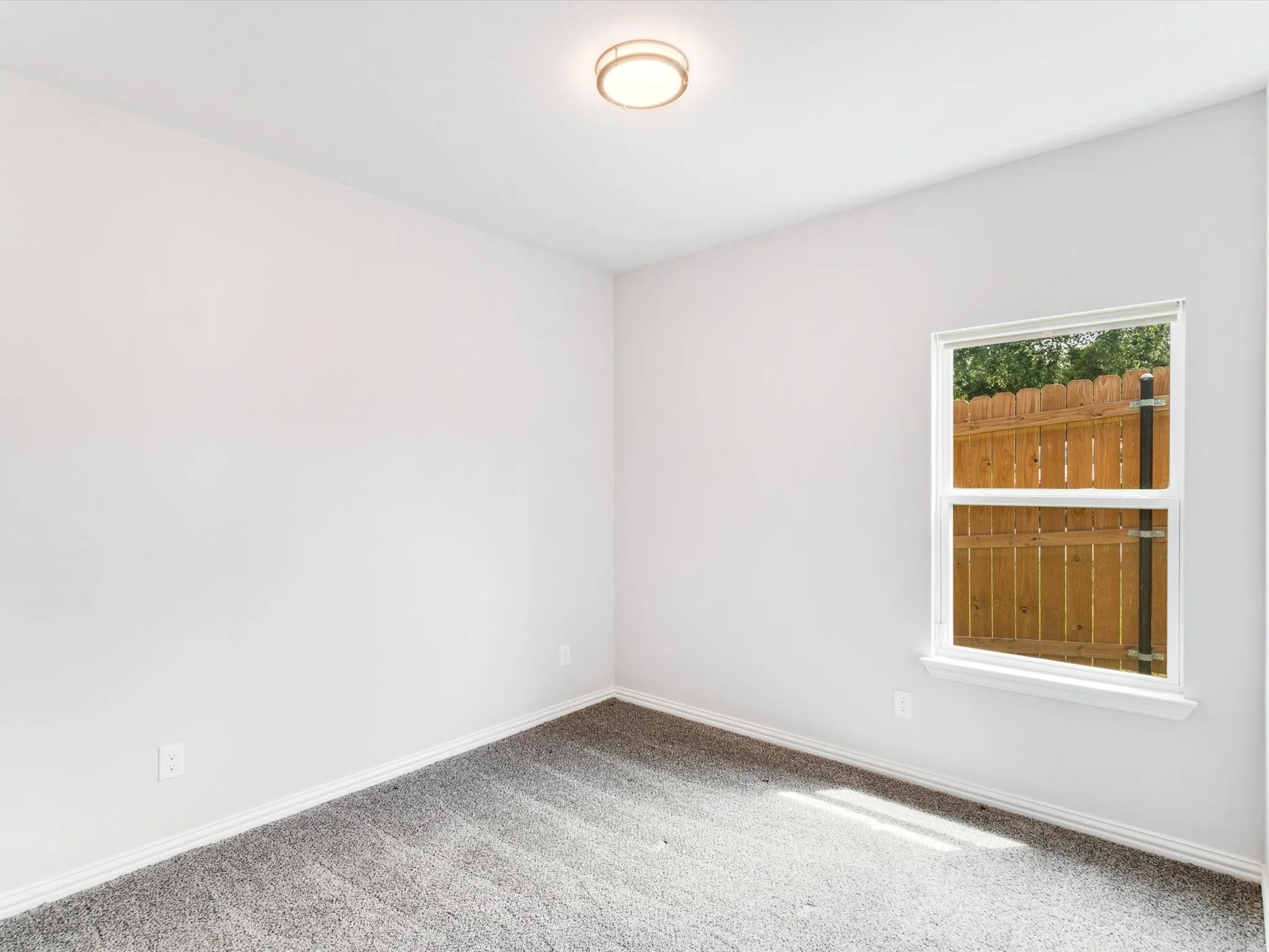 Bright interior room featuring a single window, overhead flush-mount lighting, light gray wall paint, white baseboards, and gray textured carpeting