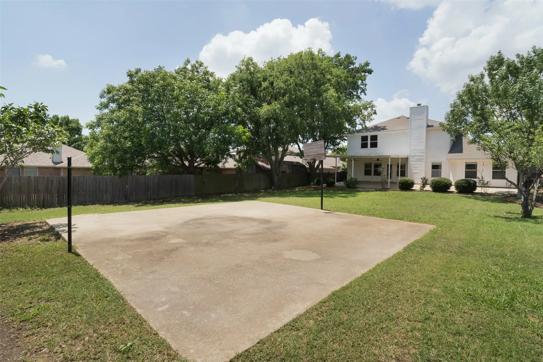 Expansive backyard featuring a large concrete court, mature trees, a wooden privacy fence, and a white two-story home with a covered patio