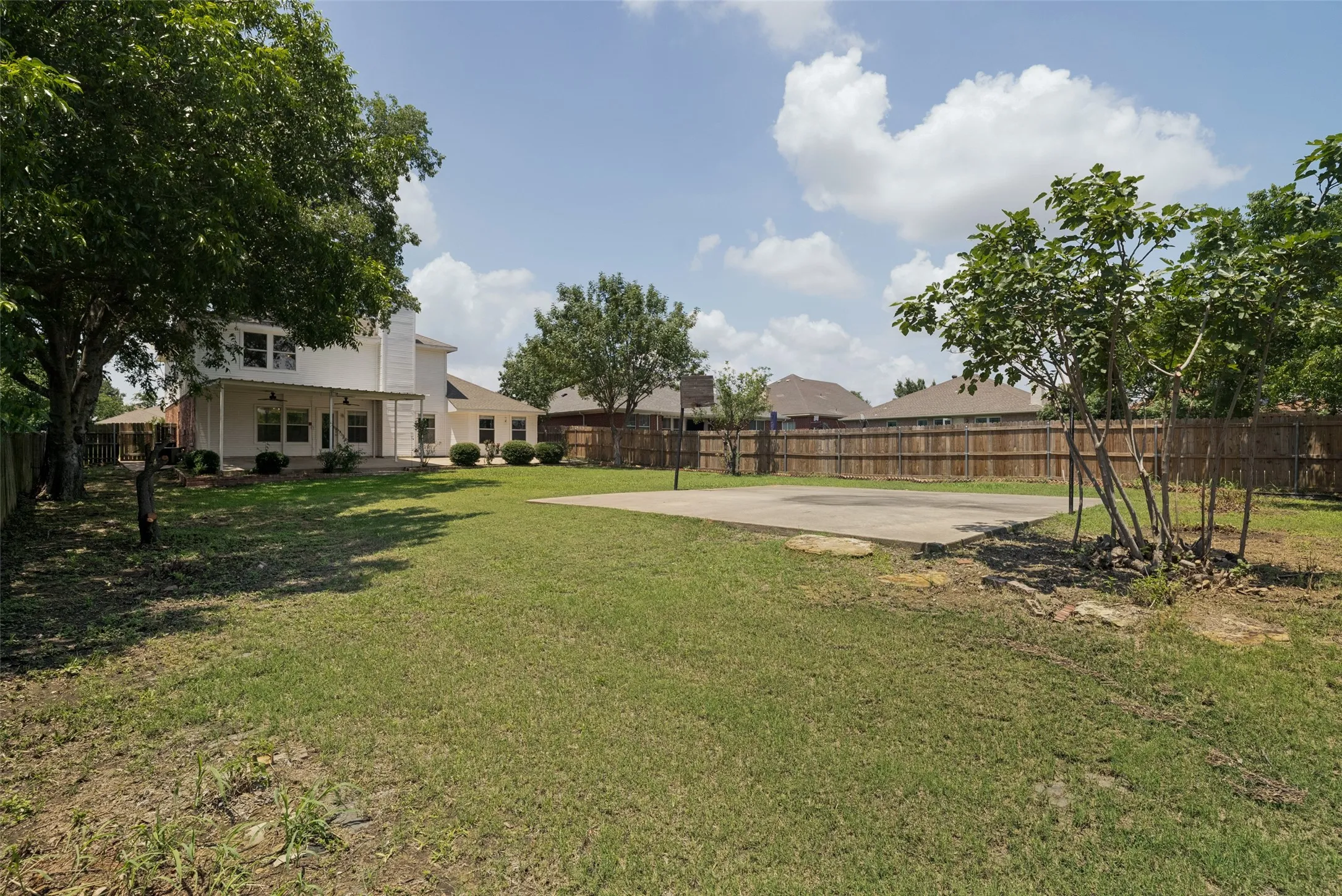 Spacious backyard featuring a manicured lawn, a large concrete pad, established trees, and a wooden privacy fence