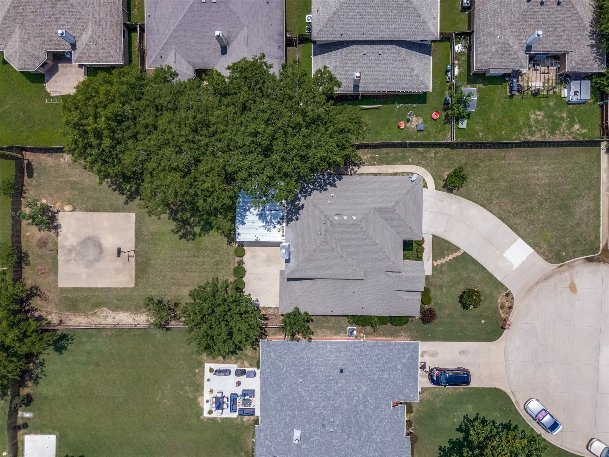 Aerial property view featuring a large concrete driveway, a private fenced yard, mature trees, and a concrete sport court