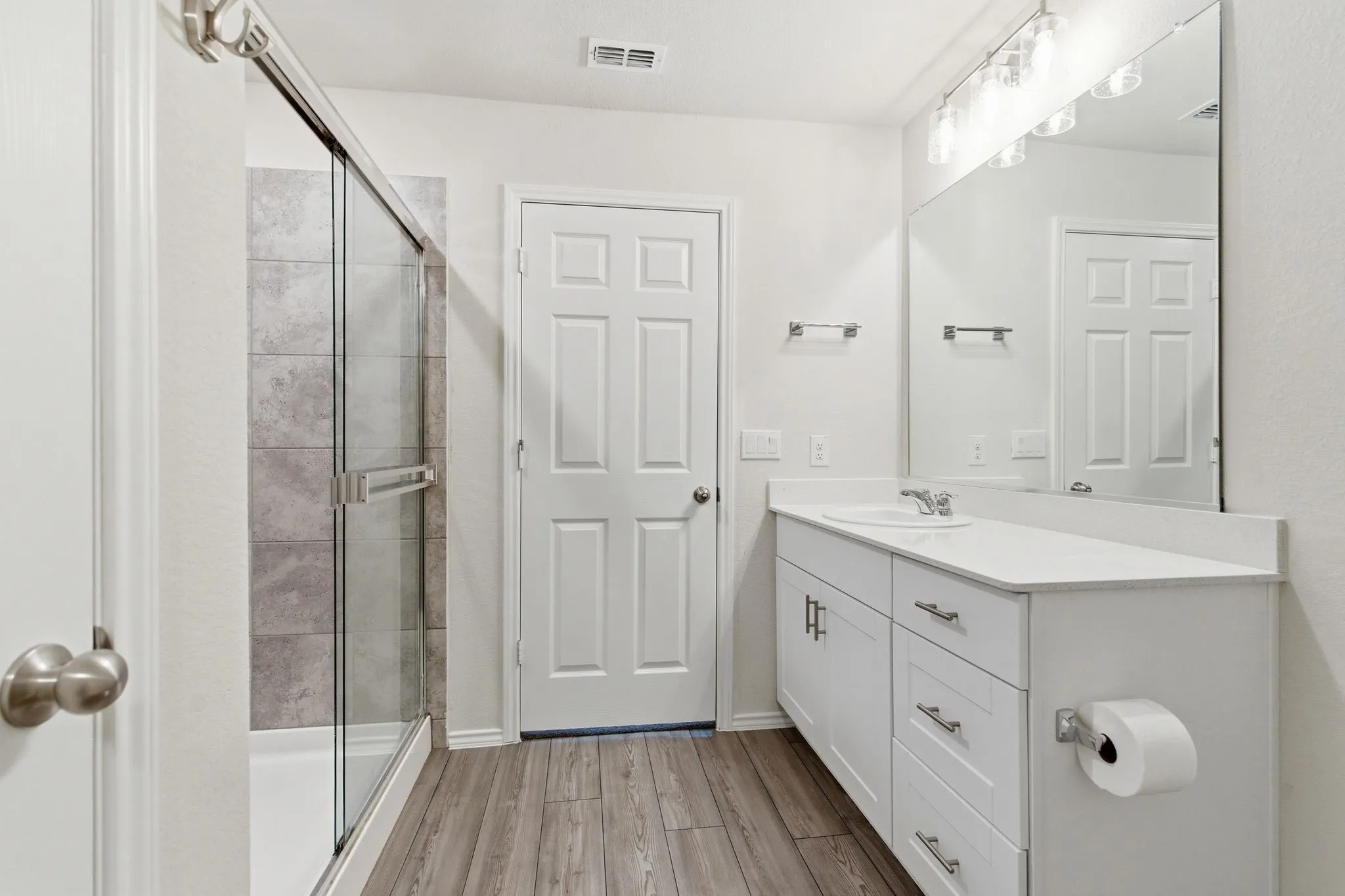 Bathroom featuring a white silestone quartz vanity with a single basin sink, a framed mirror, a glass-enclosed shower with gray tiled walls, and wood-look tile flooring